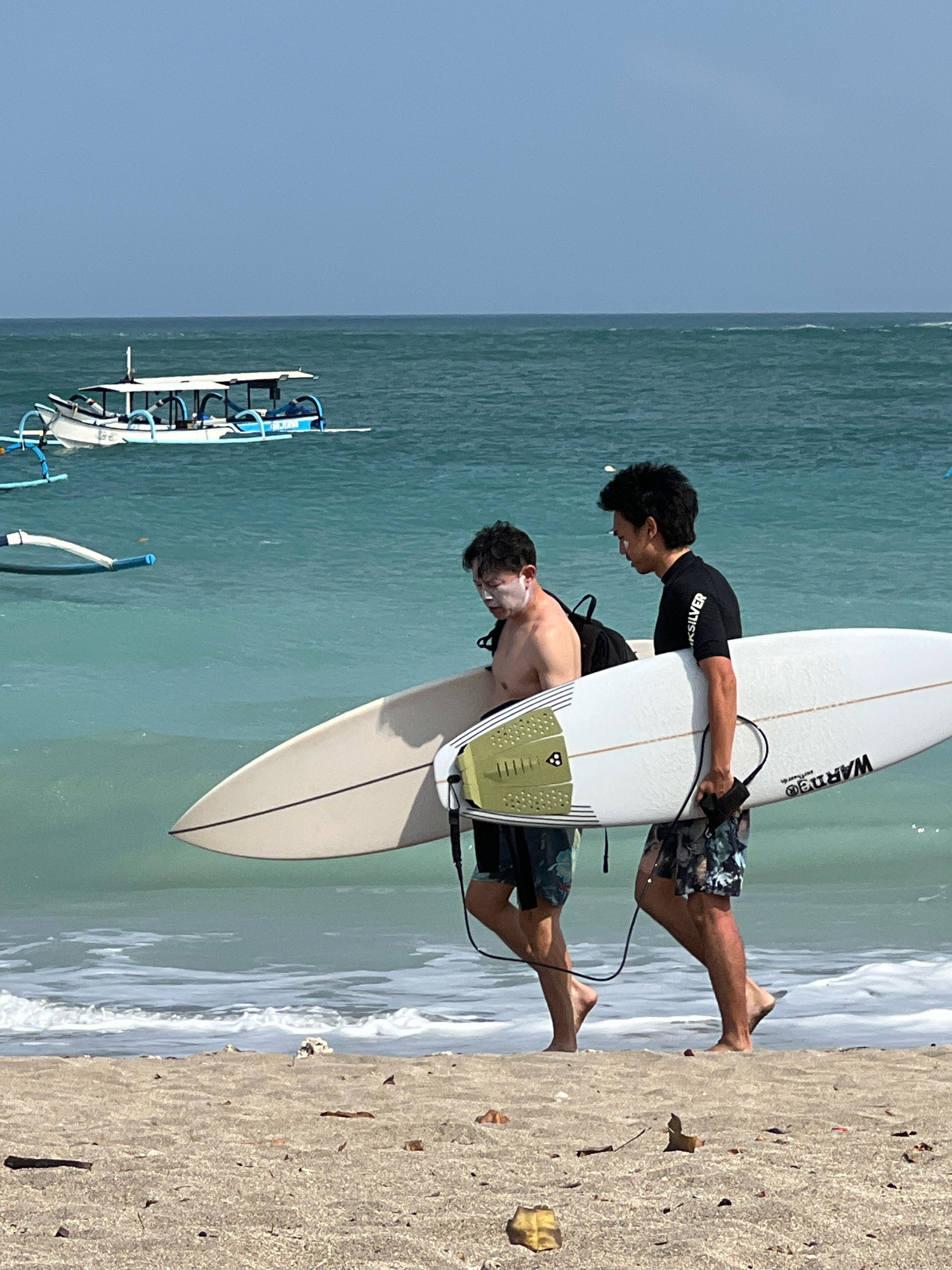 Two men are walking on a sandy beach in Kuta, Indonesia, carrying a surfboard. The man on the left, who is shirtless, has sunscreen applied to his face. The man on the right, wearing a black t-shirt with "QUICKSILVER" printed on it, carries the surfboard with a black leash attached. Both men are wearing patterned shorts. The ocean is visible behind them with gentle waves and a boat with a white canopy is moored in the distance. The sky is clear and a light blue. The sand on the beach is light brown and appears to have some debris scattered across it.