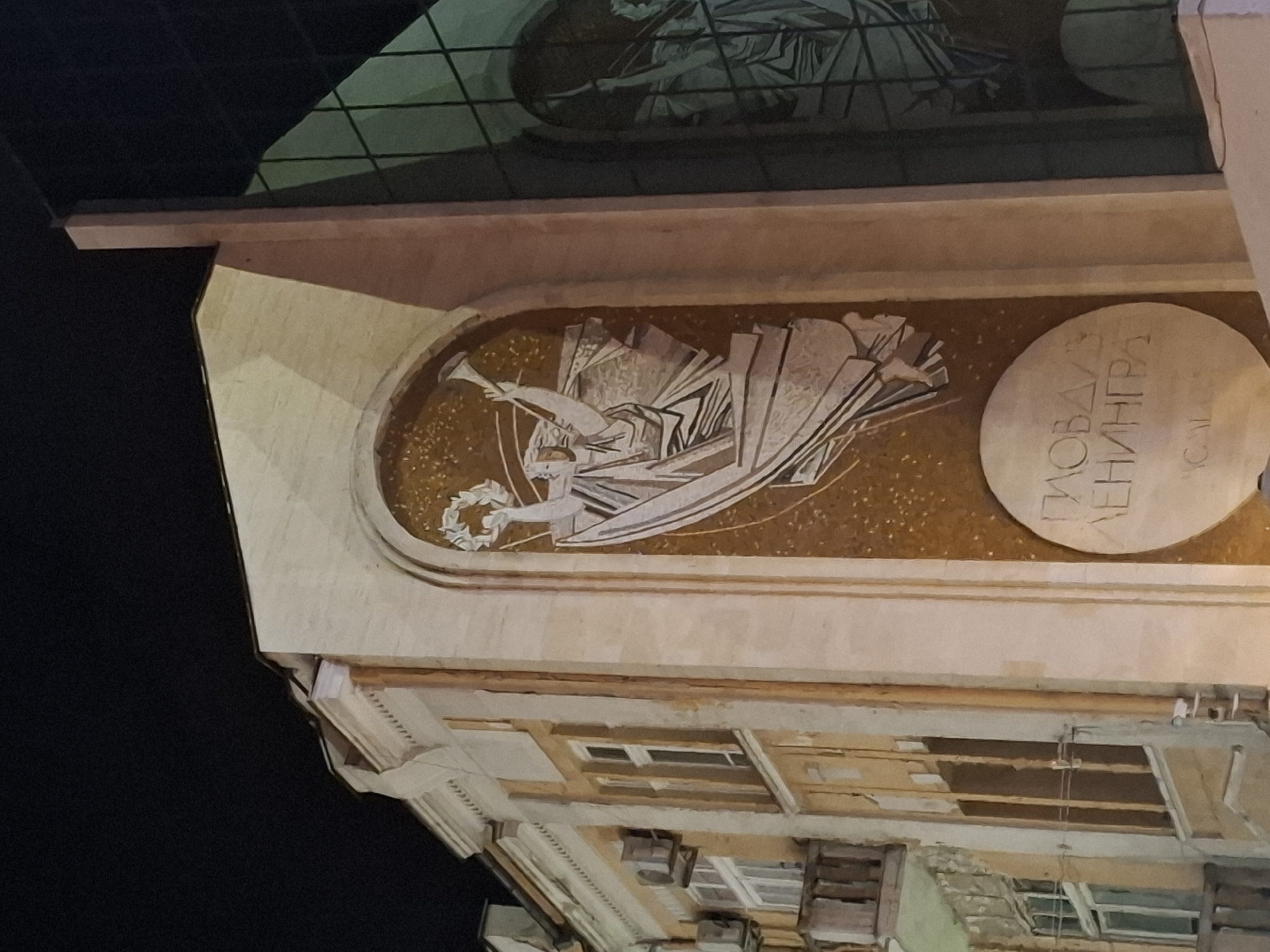 The image shows a mosaic on the side of a building. The mosaic depicts a winged figure holding a wreath and a trumpet. Below the figure, there is a circular inscription. To the left of the mosaic is a building with windows and air conditioning units. To the right is a modern building with large green-tinted windows. The scene appears to be illuminated at night.