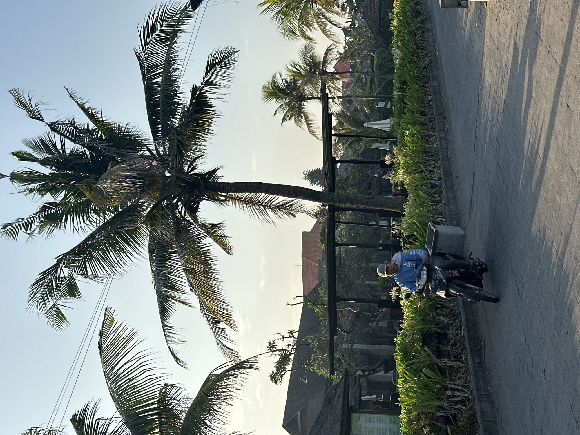 In North Kuta, Indonesia, a man wearing a blue hooded sweatshirt and a gray baseball cap rides a black motorcycle on a paved road. He is seen from the chest up, looking forward. The motorcycle has a license plate that reads "DK 1407 US" and a gray bin attached to the rear. The background features lush green foliage, several palm trees, buildings with dark roofs, and white umbrellas. The sky is a clear, pale blue, suggesting it is daytime. The overall scene depicts a tropical, possibly resort, environment.