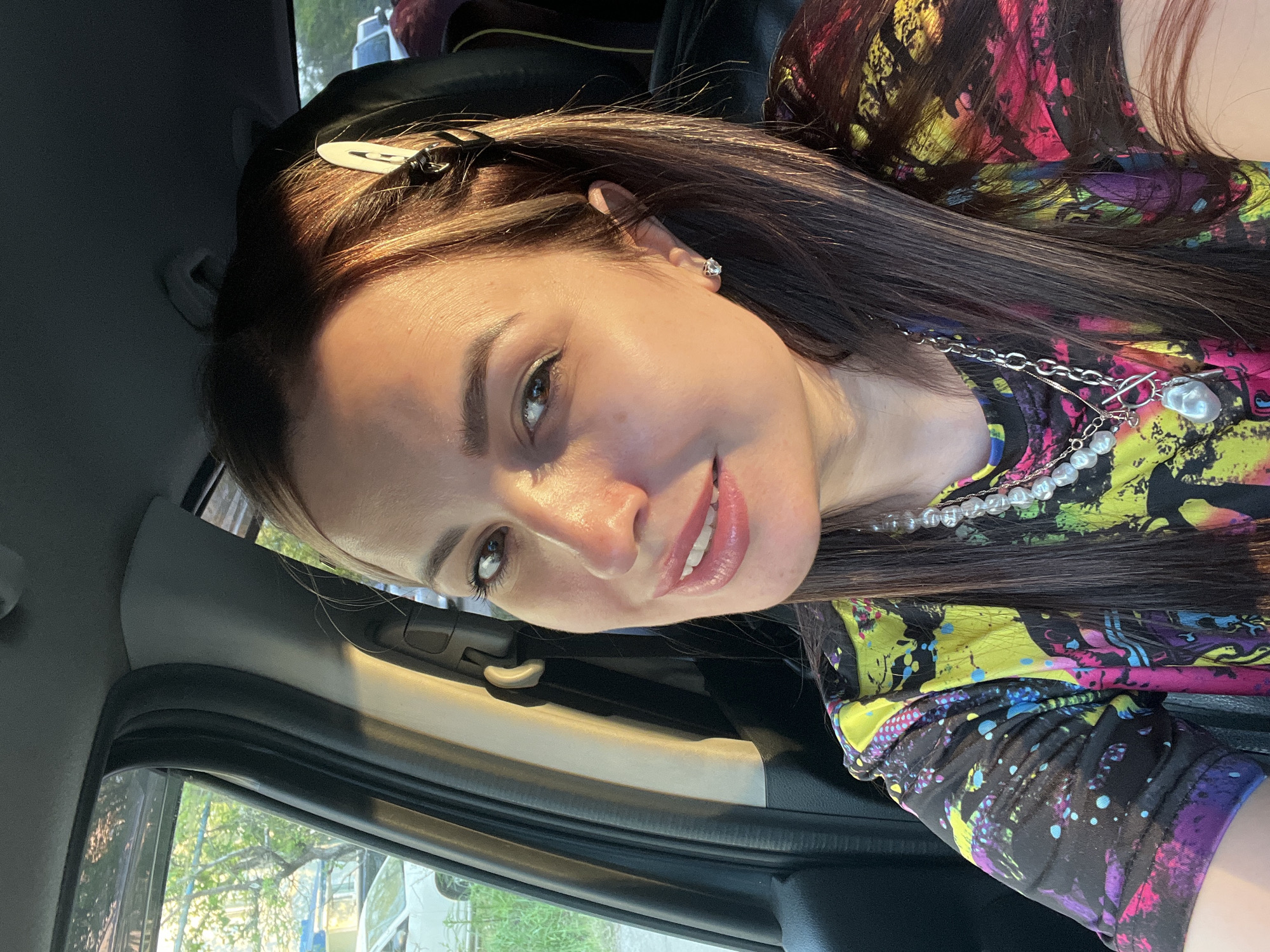 A young woman with long, dark brown hair is posing for a selfie in the front seat of a car. Her hair is styled with a white clip near her temple. She is wearing a colorful, patterned shirt and a pearl necklace with a silver chain. The car's interior is visible, including the dashboard and window frame. Outside the window, there is a glimpse of greenery, possibly trees or bushes. The location is Unknown, Unknown.