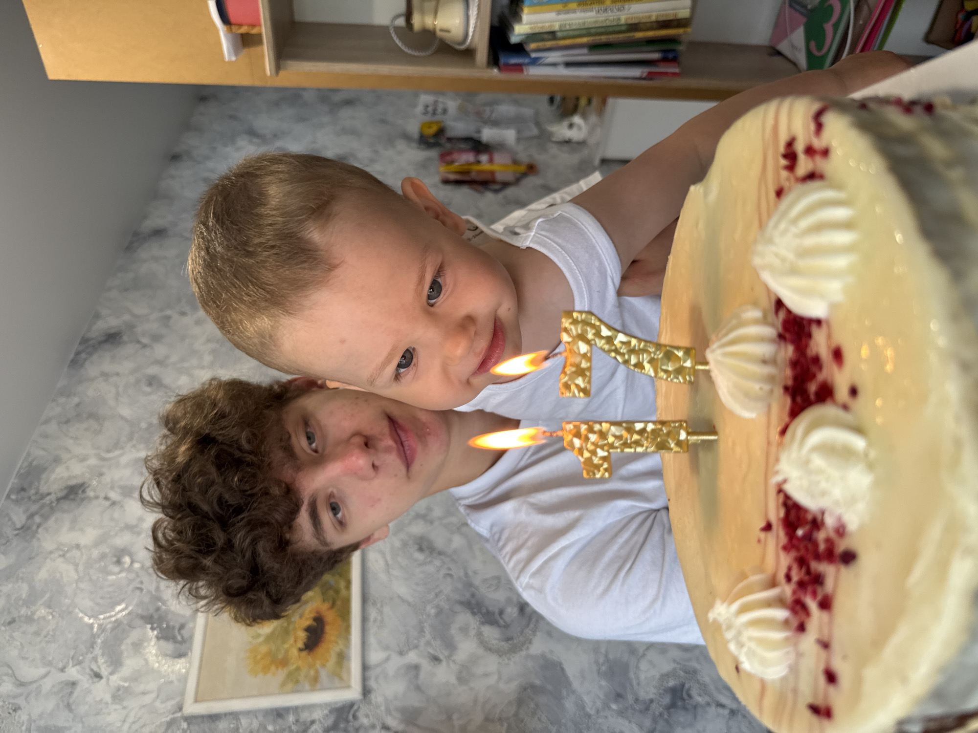 Two individuals, a toddler and a young adult, are positioned close to each other, possibly posing for a photo. Both are wearing white shirts. The toddler is on the upper side of the frame, looking toward the cake. The young adult is beneath the toddler, facing the camera with a slight smile. In front of them is a cake decorated with cream swirls and red sprinkles, with candles in the shape of the number "17" inserted on top. The candles are lit. The background features a gray marbled wall, a framed sunflower painting, and a shelf with books and other objects. The location appears to be indoors, likely a private residence. There is no information in the image indicating the location is Rusanovo, Russia.