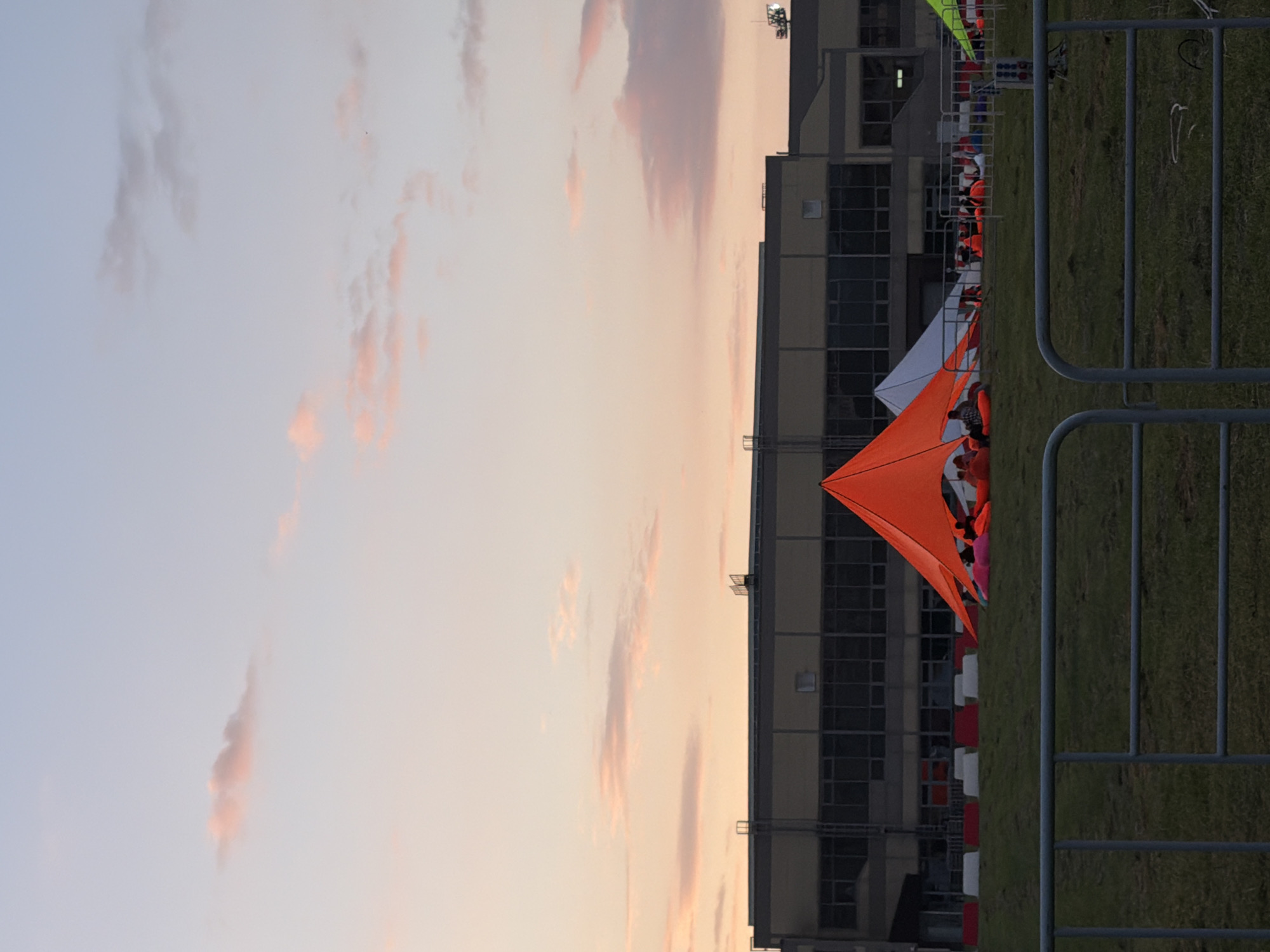The image depicts an outdoor scene with a large, modern building in the background, likely a convention center or industrial facility. In front of the building, a grassy field is set up with several tents. A prominent orange tent is in the foreground, with people seated inside and around it. Adjacent to the orange tent, a white tent is visible. The sky above is a soft gradient of pastel colors, suggesting either sunrise or sunset. A metal fence runs across the bottom of the image, indicating a boundary or viewing area. The overall atmosphere is calm and serene, with a hint of festival or gathering.