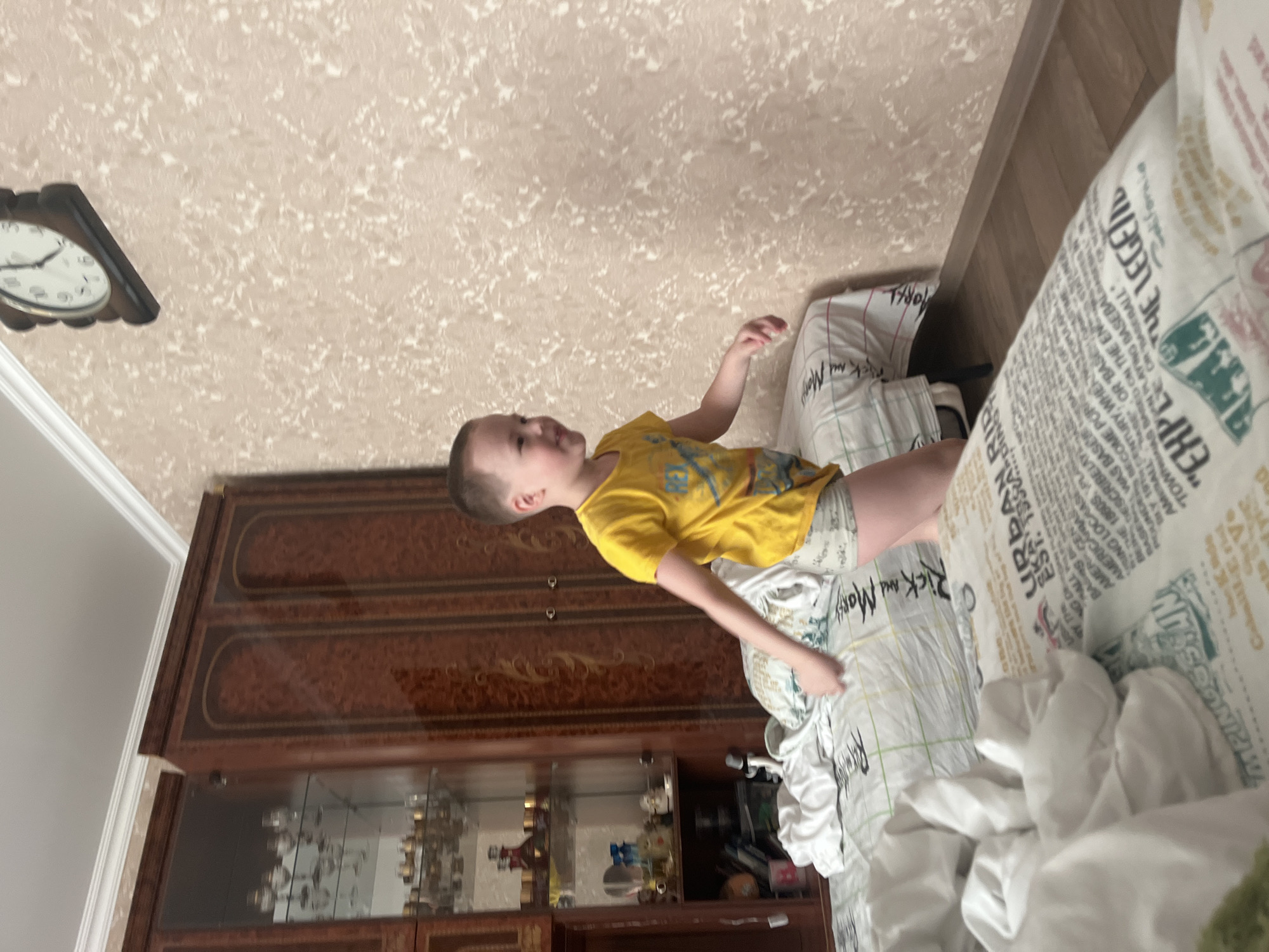 A child in a yellow shirt lies on a bed in a room. The bed has bedding with printed text. There is a cabinet with glass doors filled with items against the wall. A clock hangs on the wall. The specific location in Usman, Russia, is not discernible from the image.