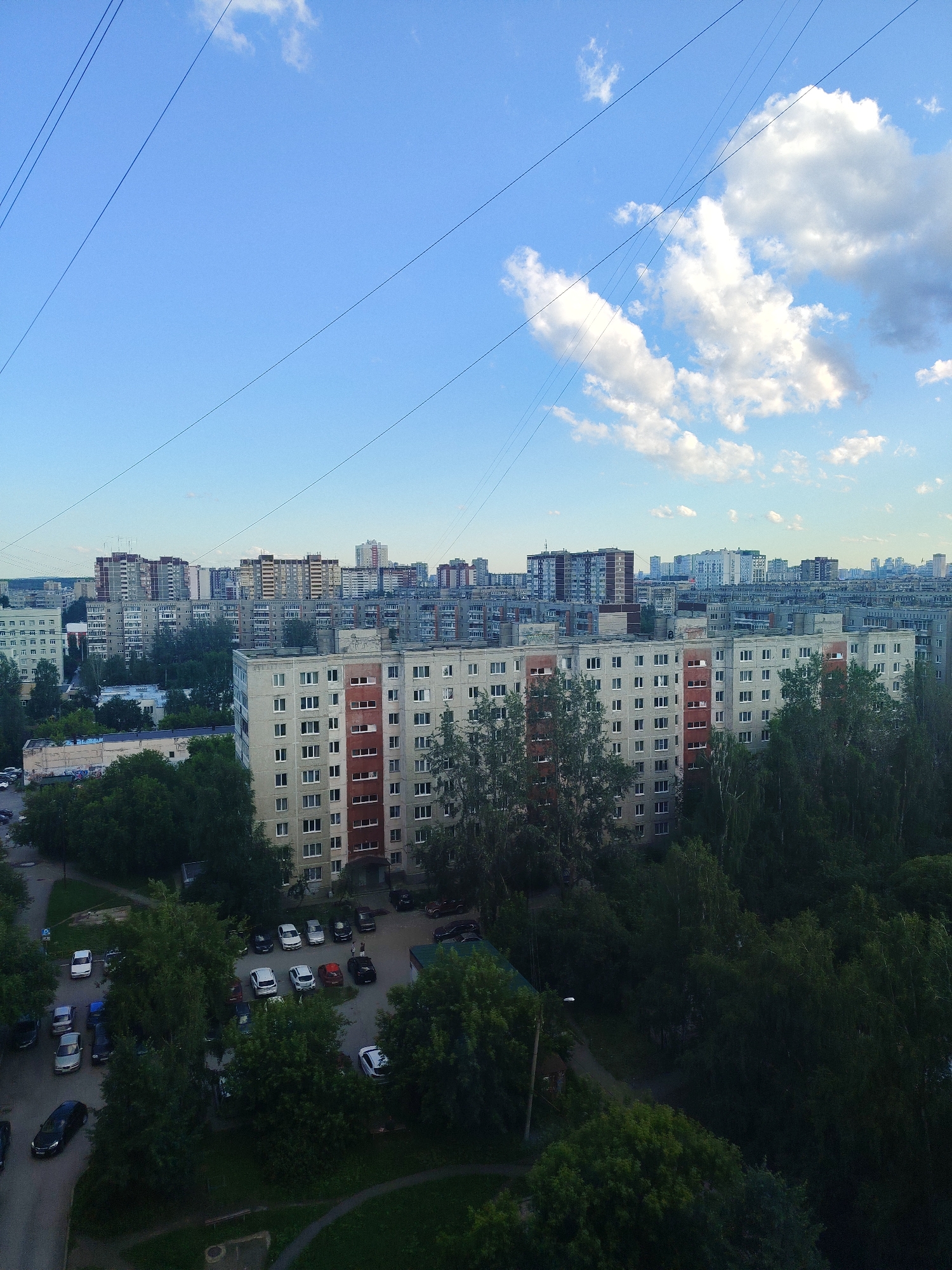 This is an aerial view of a residential neighborhood in Yekaterinburg, Russia, on a partly cloudy day. The scene depicts a dense urban landscape dominated by apartment buildings of varying heights and architectural styles, typical of Soviet-era construction. The buildings are predominantly light-colored, with some featuring red-brick accents.
In the foreground, a large, multi-story apartment building with many windows stands out. Beyond it, a dense canopy of green trees obscures the lower sections of other buildings and forms a significant part of the natural landscape within the urban setting. Numerous cars are parked in open areas between buildings and on designated parking lots, suggesting a populated area. Some of these cars are visible in detail in the lower left and center of the image.
Overhead, the sky is a clear blue, punctuated by fluffy white cumulus clouds. Several power lines or communication cables crisscross the sky, a common sight in urban environments. The lighting suggests it is either late afternoon or early morning, given the long shadows and the quality of light on the buildings and clouds. There are no visible people in the frame, and no text is discernible. The overall impression is of a typical, functional, and somewhat monotonous urban environment, with the natural elements of trees and sky providing a contrast to the man-made structures.