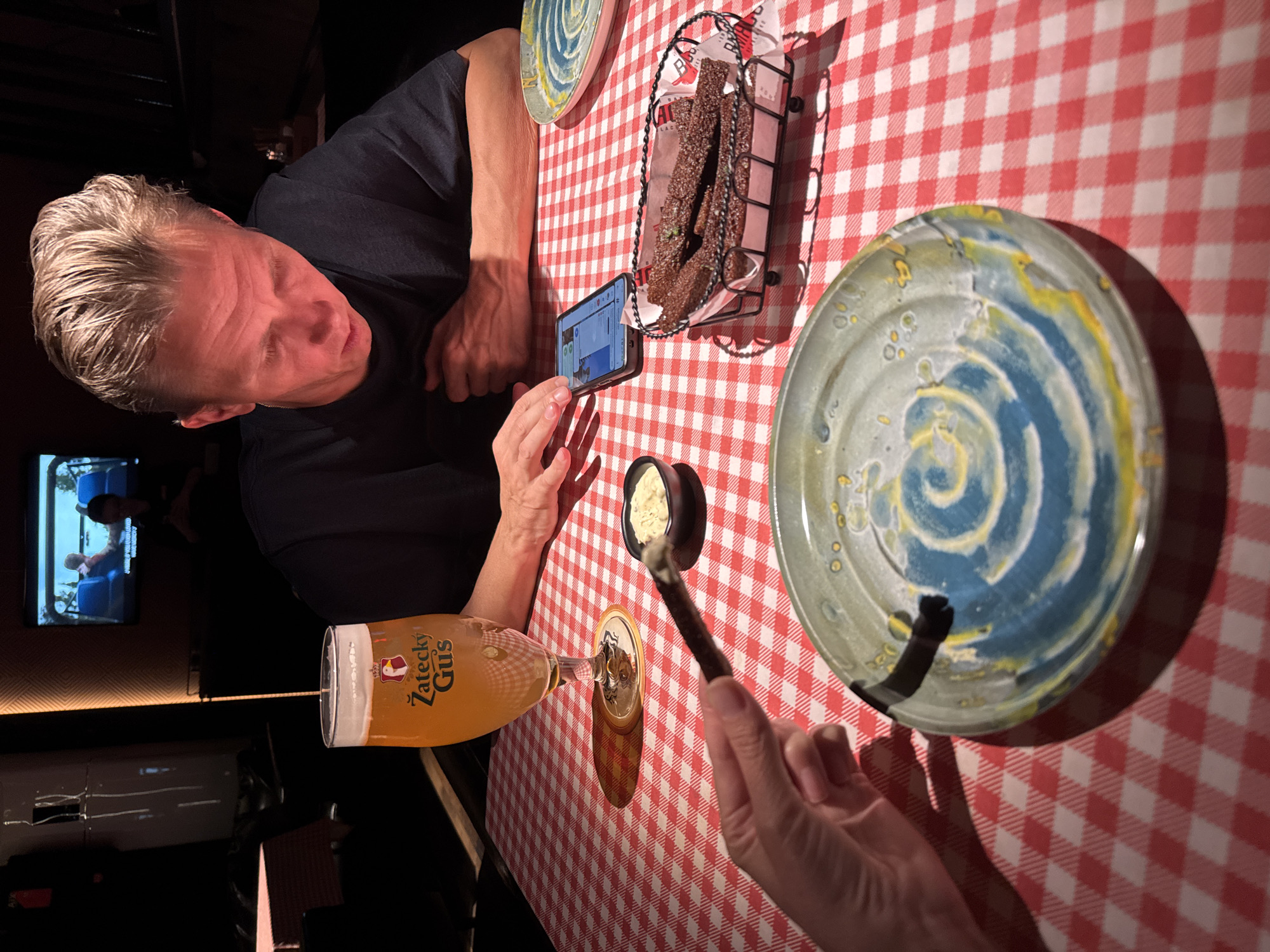 A restaurant scene in Tashkent, Uzbekistan, shows a table covered with a red and white checkered tablecloth. A man with short, light-colored hair is seated and looking at his cell phone. On the table are several plates, a basket of dark breadsticks, a small container of dip, and a glass of "Zatecky Gus" beer. Someone's hand holds a breadstick, apparently about to dip it. A television is visible in the background, suggesting the restaurant has a bar or entertainment area.