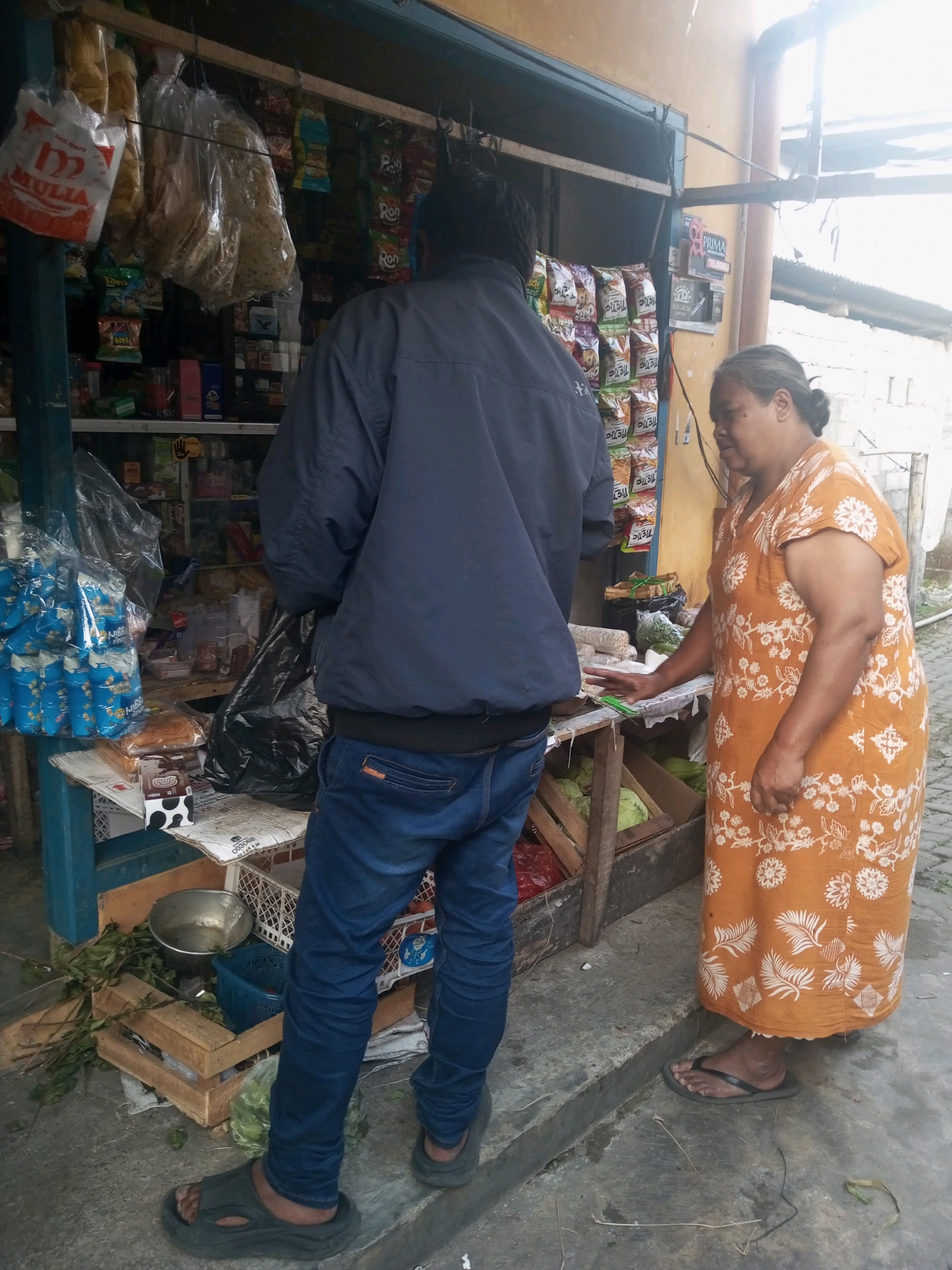 An over-the-shoulder, medium shot captures a man in a dark blue jacket and blue jeans standing at a market stall. He faces away from the camera, toward the stall where a woman in an orange floral dress stands. The woman is looking at the man and reaching towards the stall with her right hand. The stall is filled with various packaged goods, snacks, and some produce. Behind the man, various products are displayed on shelves and hung from the ceiling, including bags of snacks and drinks. The ground in front of the stall is concrete, and there are some wooden crates and greenery scattered around. The overall scene suggests a transaction or interaction at a small roadside market in Andongsili, Indonesia.