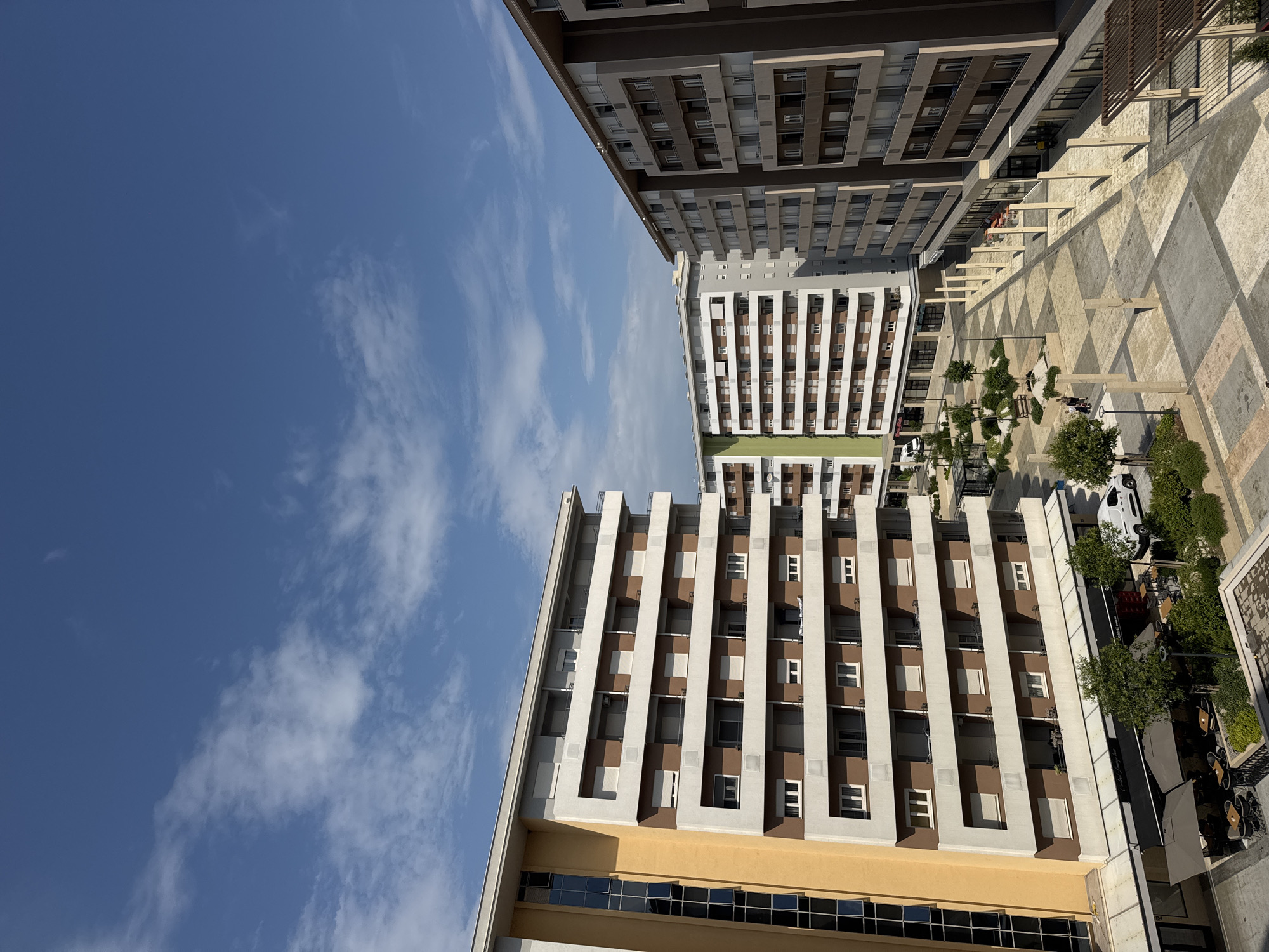 An eye-level, medium shot captures a modern urban courtyard flanked by multi-story apartment buildings under a bright blue sky with scattered white clouds. The building on the left is predominantly yellow and features a vertical strip of dark windows. Adjacent to it, and extending into the midground, is a building with a facade of alternating brown and white horizontal bands, each level punctuated by balconies and windows. To the right of this, another similarly structured apartment building with brown and white sections is visible. In the foreground, the courtyard features a checkered pattern of light-colored paving stones, with trees, seating areas, and a white van. In the background, more buildings and a trellised structure are observed. The scene depicts a residential area on a clear day.