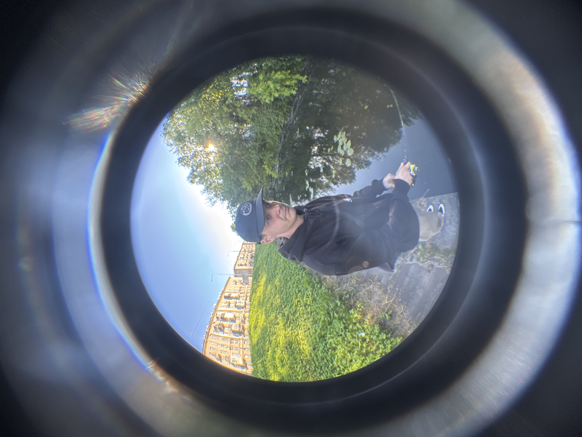 A man wearing a black hoodie and a baseball cap is fishing by a body of water in Saint Petersburg, Russia. The image is captured with a fisheye lens, creating a distorted, circular view. The man is holding a fishing rod and appears to be casting. The background shows a grassy embankment leading up to a building and trees. The sun is visible through the trees, casting light on the scene. The water has lily pads floating on its surface. The scene is framed by a dark, circular border, characteristic of a peephole or a fisheye lens.