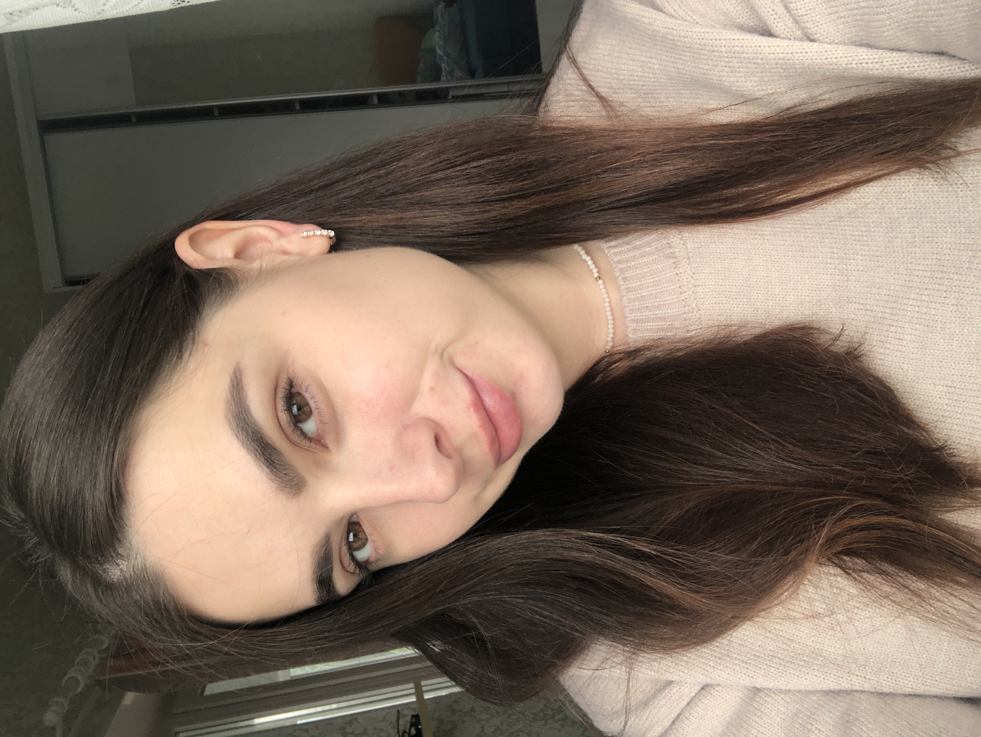 A portrait-style photograph features a young woman with long, dark brown hair, looking directly at the viewer with a slight, gentle smile. Her head is tilted to her right. She has brown eyes and well-defined, dark eyebrows. Her skin appears clear, and her lips are naturally pink.
She is dressed in a light-colored, possibly beige or light pink, knitted sweater. Around her neck, she wears a delicate choker or necklace made of small, light-colored beads. On her left ear, she has a small ear cuff or earring embellished with a line of clear, sparkling stones. Her long hair flows down, covering parts of her shoulder and the sweater.
The background is dark and out of focus, indicating an indoor setting. Indistinct shapes are visible in the blurred background, and there's a hint of a lighter, possibly lacy, material at the top, which might be a curtain. The lighting is soft and even, illuminating her face. The overall mood of the image is calm and inviting. There is no visible text in the image. The location context for the image is Noviy, Russia.