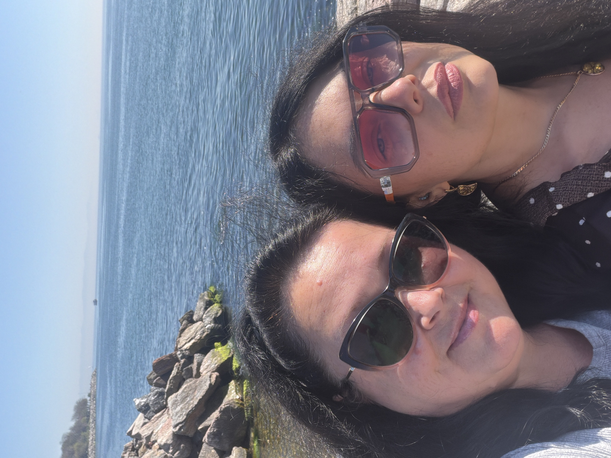 Two women are posing for a selfie outdoors, seemingly near the water. The woman at the top of the frame has dark hair, sunglasses with a red-brown tint, and is wearing a black and white polka dot top with a gold necklace. The woman at the bottom of the frame also has dark hair and sunglasses, with a lighter colored shirt. Behind them is a body of water and a rock formation. The photo was taken in Kadıköy, Turkey.