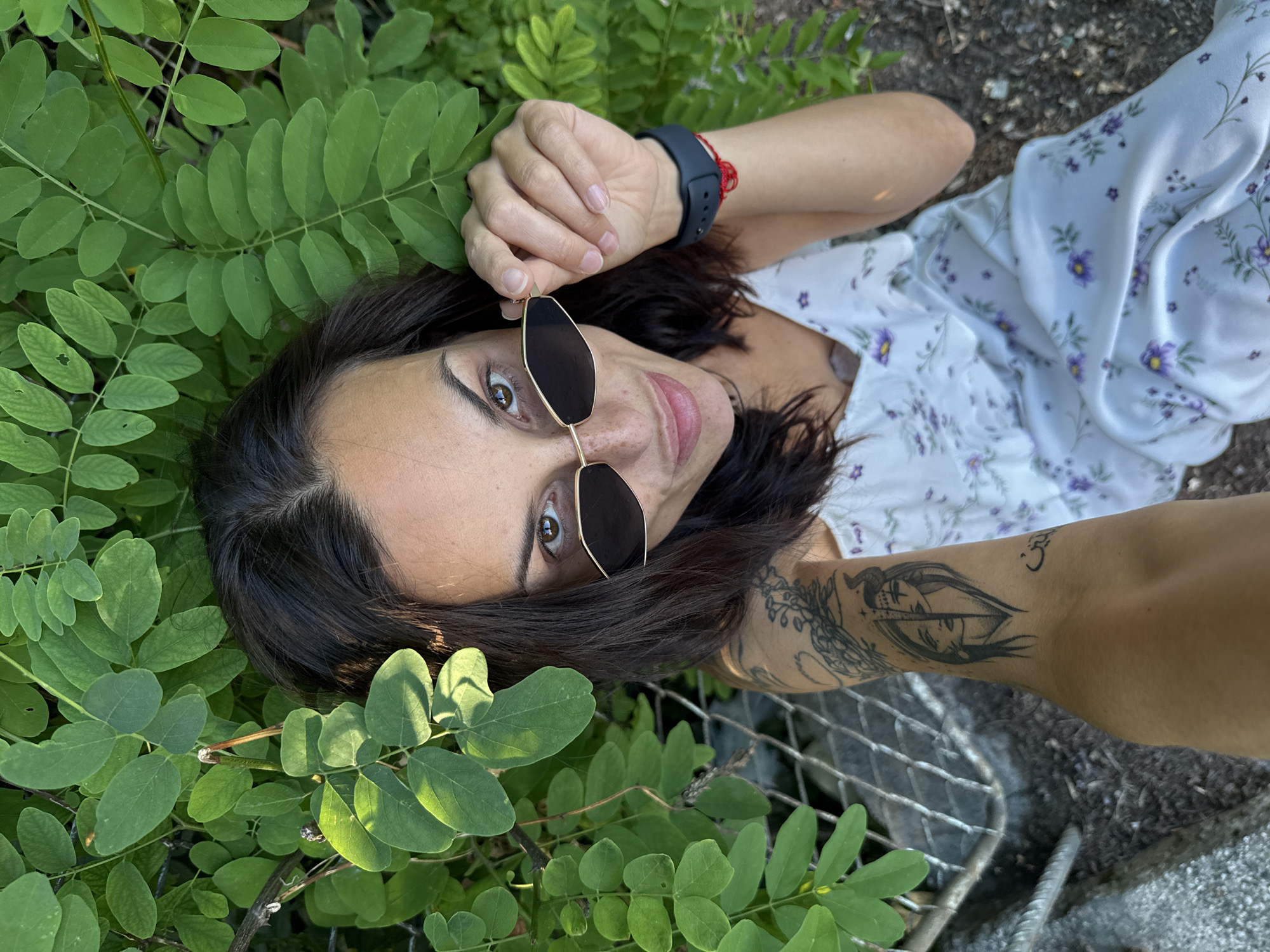 In Pleven, Bulgaria, a young woman with dark hair and brown eyes is pictured from a high-angle perspective. She is wearing a white, floral-patterned slip dress and dark-rimmed sunglasses, which she is adjusting. Her left arm, adorned with a tattoo of a half-face with horns, is extended toward the camera. She is positioned outdoors, surrounded by lush green foliage, with her arm resting on what appears to be a metal grate or fence. A dark-colored smartwatch is visible on her right wrist, with a red string bracelet tied around it. The ground beneath her is covered in soil and small stones.