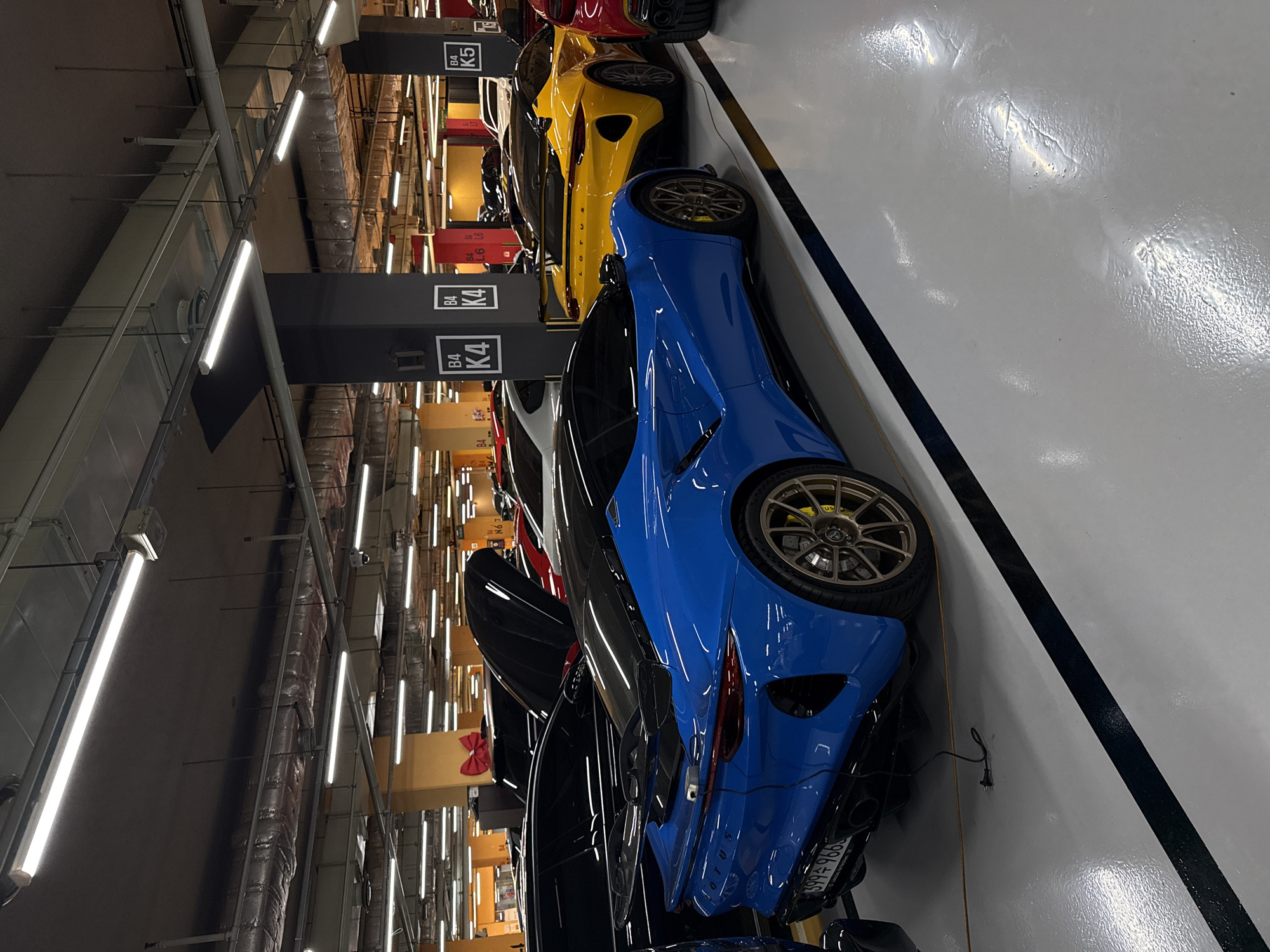 The image depicts a parking garage in Seoul, South Korea. Several luxury cars, including blue, yellow, red, white, and black Lotus models, are parked in a row. The cars are parked under a series of overhead lights and parking bay markers labeled "B4" and "K4," "B4" and "L6," and "B4" and "K5." The parking space is marked with a black stripe along the edge. The floor is a light grey color and reflective. The license plate of the blue Lotus is visible.
