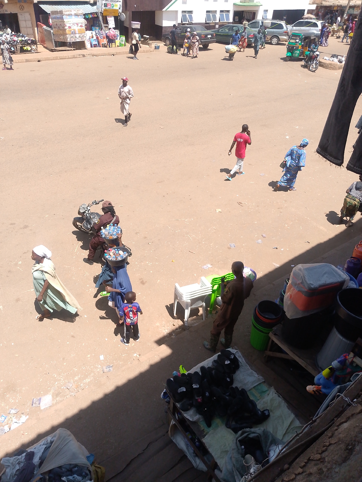 The overhead shot captures a bustling marketplace in Bukuru, Nigeria, bathed in bright sunlight. The ground is a mix of dirt and some pavement, with scattered debris. In the foreground, a pile of folded dark clothing rests on a mat, and further back, more items are stacked on shelves.
People are actively moving through the scene. A man in a white robe and red cap walks from right to left. To his left, a woman in a pale green garment and white headscarf walks towards the foreground. Behind her, a child in a red and blue jersey with the number 9 on the back walks forward. Two women, one in a blue patterned dress and the other in a darker blue dress, are walking together carrying baskets. One of the women appears to be pushing a cart with items. In the background, a man in a red shirt is seen talking on his phone. Further back, a green and yellow tricycle is parked. Several vehicles, including cars and a motorcycle, are visible on the street. Market stalls and buildings line the background, with awnings and various goods displayed. The overall impression is one of daily commerce and activity in a Nigerian urban environment.