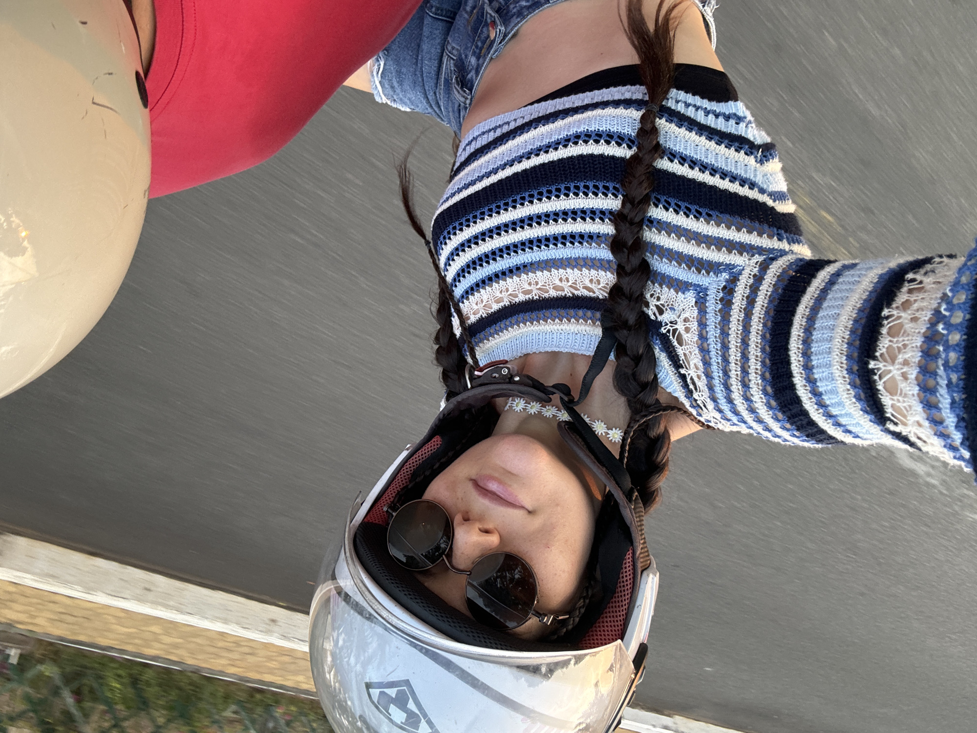 The image is a close-up, low-angle selfie of a young woman on a motorcycle, likely in Phu Thuy, Vietnam. She is wearing a shiny silver and white full-face helmet with the visor pushed up, revealing her face. Dark, round sunglasses cover her eyes, and her lips are slightly parted in a relaxed or serene expression. Around her neck, she wears a delicate choker necklace adorned with small white daisy-like flowers.
Her long, dark hair is styled into two thick braids that fall over her shoulders. She is dressed in a blue, white, and dark blue striped knitted crop top, which reveals her midriff, and light blue denim shorts. To the far left of the frame, part of another person or object is visible, featuring a red garment and a light-colored helmet.
The background appears blurred, suggesting motion, and depicts a paved road or highway with a hint of green foliage along the bottom edge, indicative of a roadside environment. The bright, even lighting points to a daytime setting. A partial logo, possibly including stylized letters like 'K' and 'Y', is visible on the side of the woman's helmet. The image conveys a casual moment during a journey.