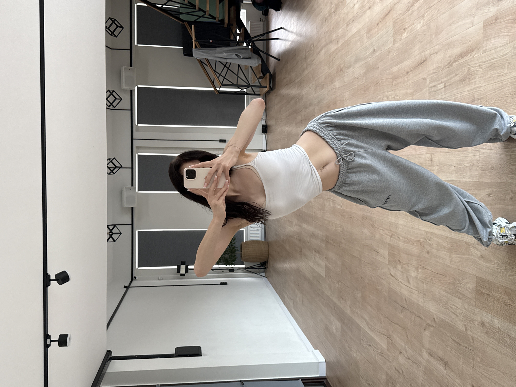 In a dance studio with light wood floors and white walls, a young woman with dark hair takes a mirror selfie. She is wearing a white tank top, oversized grey sweatpants with drawstrings at the waist, and athletic shoes. The studio is illuminated by track lighting on the ceiling. The background shows windows with dark blinds, a potted plant, and a metal staircase.