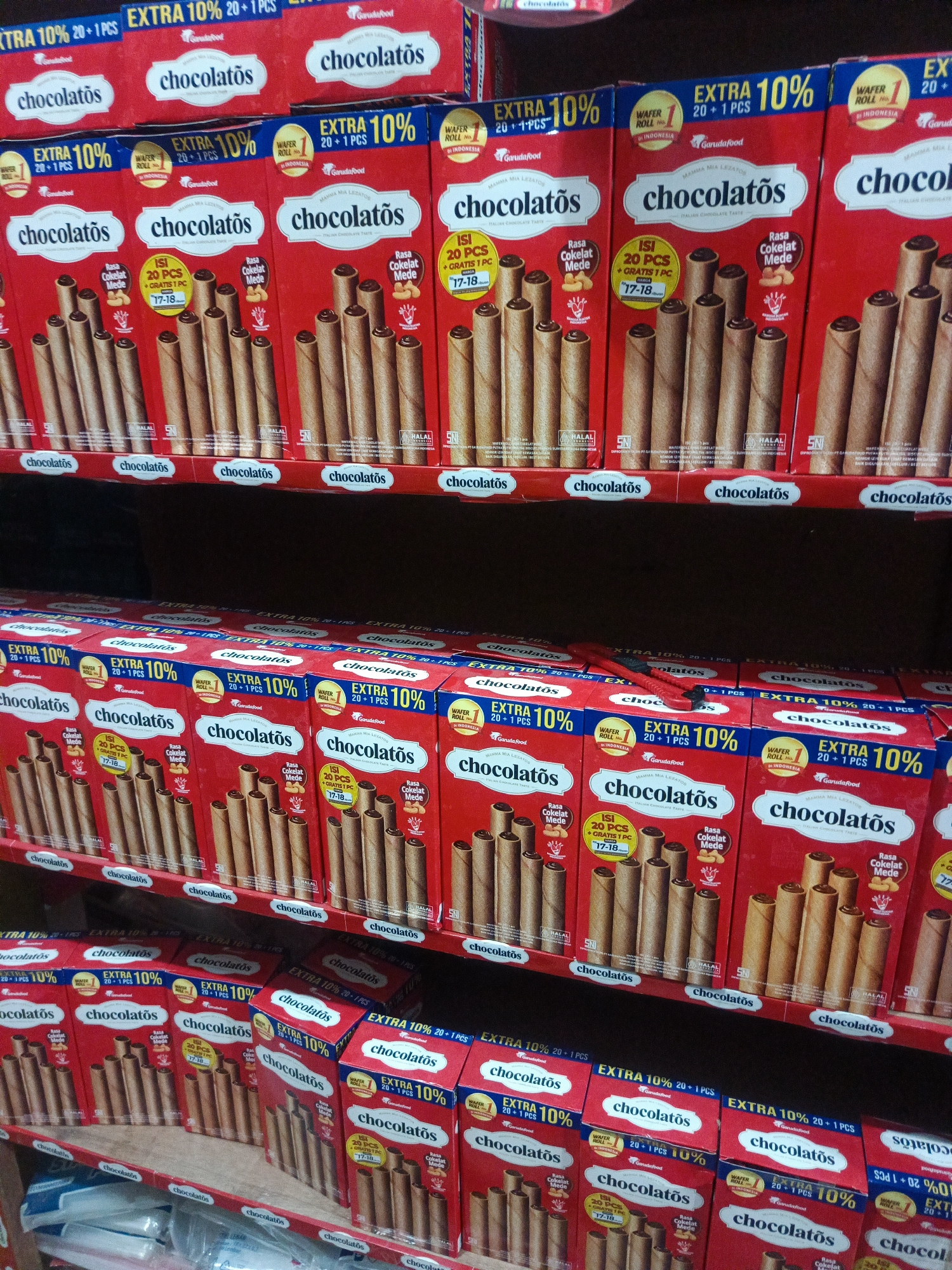 The image displays shelves filled with boxes of "Chocolatos," a brand of chocolate wafer rolls, likely in a retail store in Ilotidea, Indonesia. The boxes are predominantly red and feature the brand's logo and promotional text such as "EXTRA 10% 20 + 1 PCS." The wafer rolls themselves are visible through cutouts on some of the boxes, showing their cylindrical, brown appearance. The shelves are spaced and appear to be made of wood, with some darker shelving units visible above and below the primary display. The lighting suggests an indoor environment, possibly a supermarket or minimarket. No people are visible in the frame.