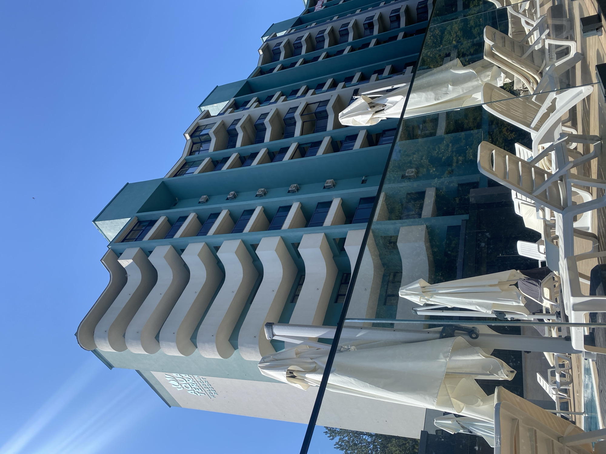 The image depicts the Aqua Terra Resort in Ustia, Moldova. The resort features a multi-story building with a distinctive turquoise and white facade, characterized by curved balconies and numerous windows. The building is viewed from a ground-level perspective, with a clear blue sky overhead. In the foreground, a swimming pool area is visible, furnished with white lounge chairs and closed umbrellas. A glass barrier separates the pool area from the viewer. The overall scene conveys a sense of a sunny day at a holiday resort.
