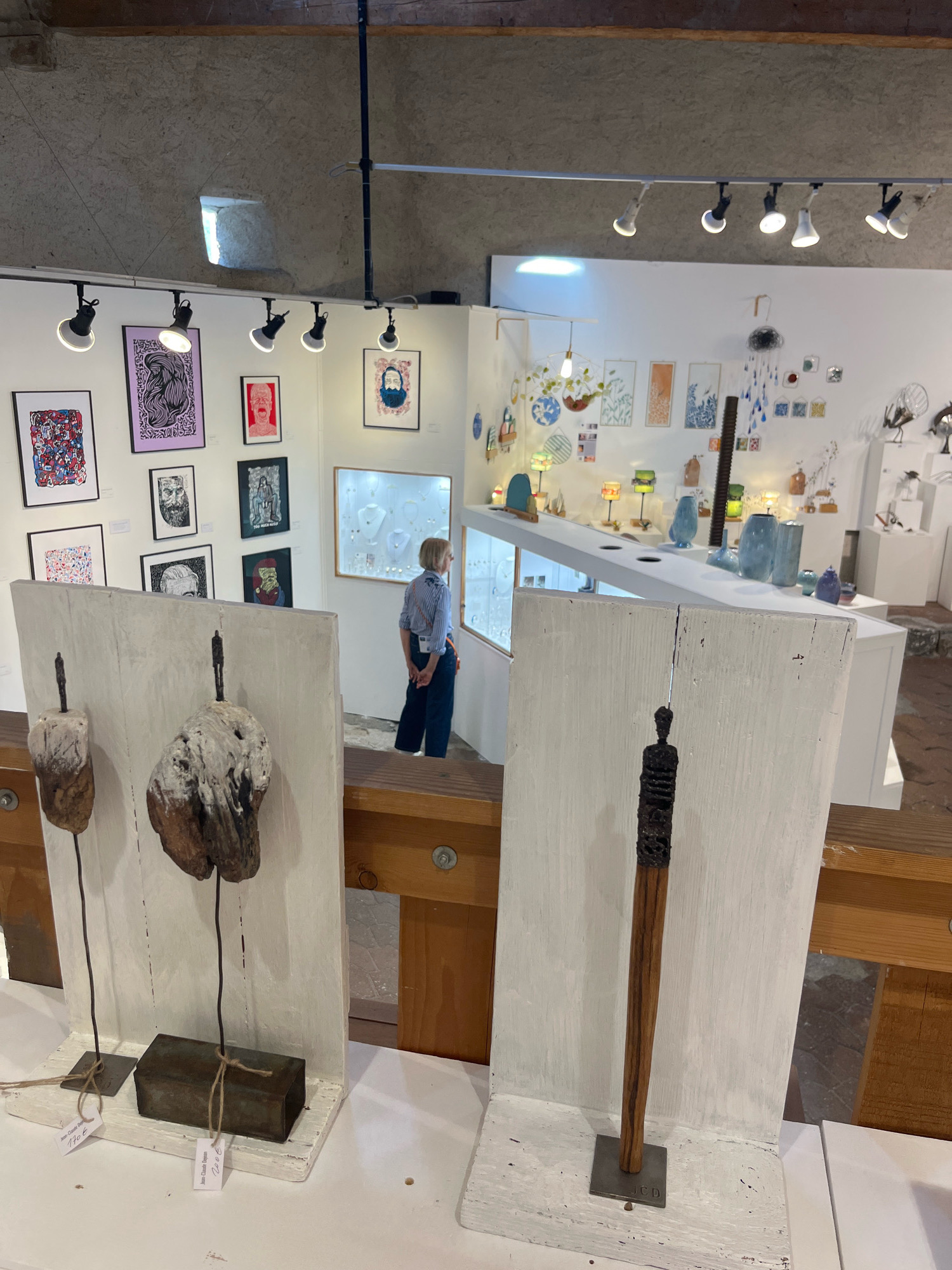 This image captures an interior view of an art gallery in Yvoire, France. In the foreground, two white wooden display stands each hold a distinct sculpture. The left stand features two abstract figures mounted on what appears to be driftwood, secured with thin metal rods and resting on metal bases. The right stand displays a single, taller wooden sculpture with a carved top, mounted on a square metal base. Both sculptures have small white tags attached.
A woman in a striped blue and white shirt and dark pants is visible in the mid-ground, standing and looking into a glass display case filled with jewelry. The display case is part of a long, white counter. Behind the counter, the gallery walls are adorned with various framed artworks, including prints and paintings. Several small lamps with colored shades are also visible on the counter.
The gallery's ceiling is high and features exposed beams and a rough, textured finish. A track lighting system illuminates the space, with several spotlights directed downwards. A small, square window with rough stonework is visible high on the left wall. The overall ambiance suggests a well-lit, curated exhibition space.