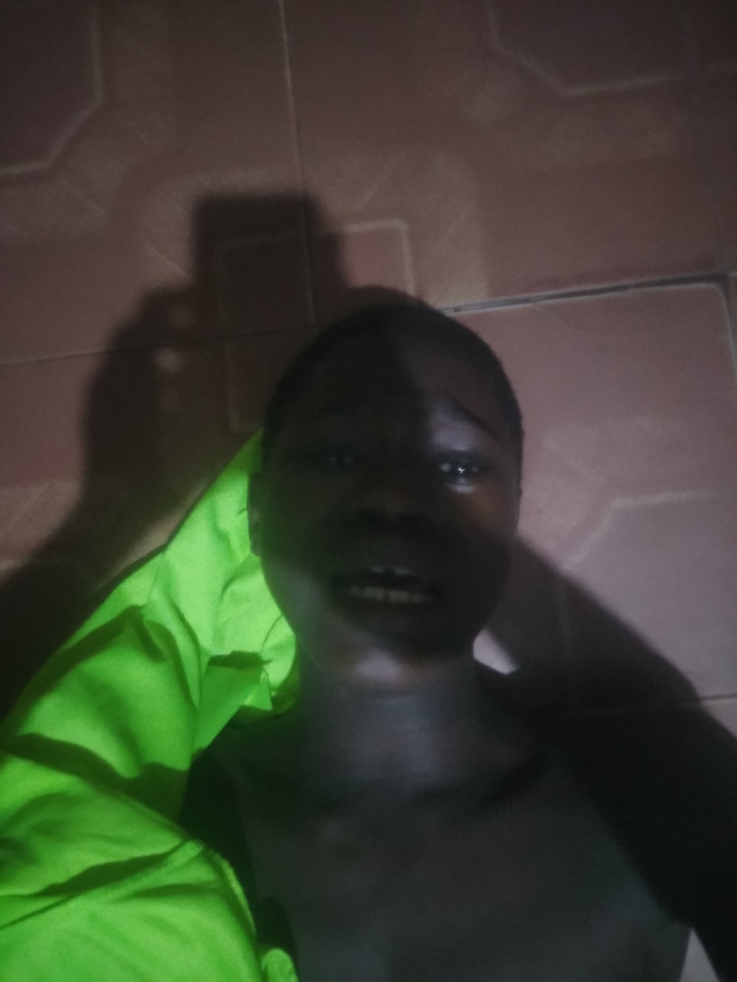 A dark-skinned person is lying on a reddish-brown tiled floor. They are facing the camera with their head and upper torso visible. They appear shirtless, and part of a bright green cloth or garment is draped over their left shoulder. A shadow suggesting the presence of a hand holding a device is cast on the wall behind them. The location is indicated to be in Shangisha, Nigeria.