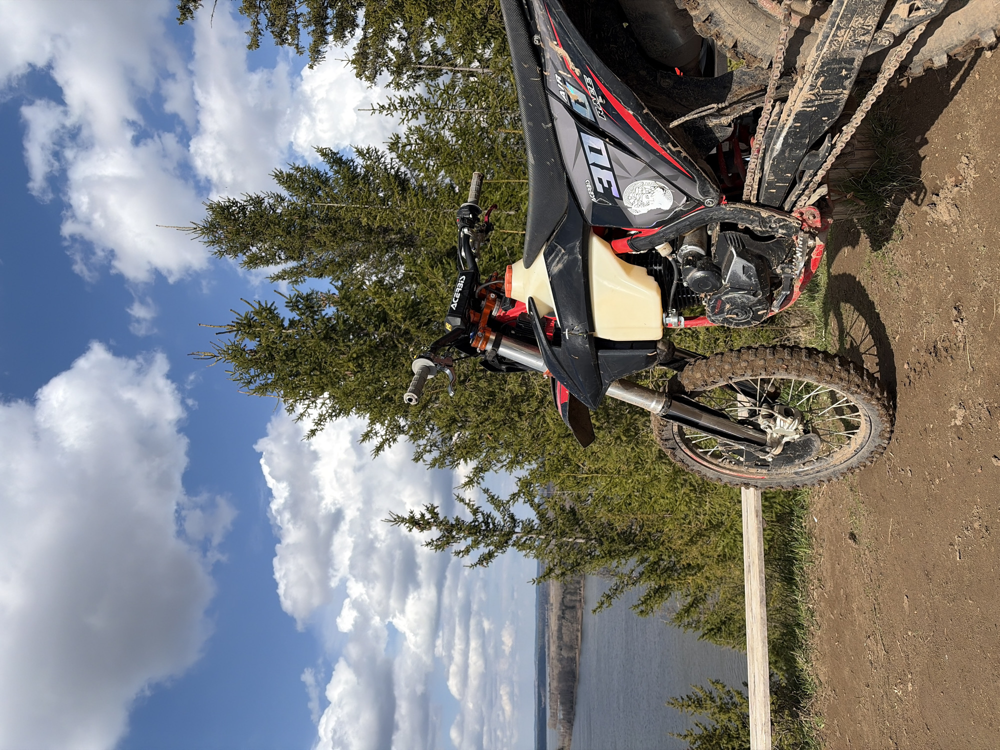 The image shows a dirt bike parked on a dirt path, with a view of a lake and trees in the background under a blue sky with white clouds. The dirt bike is black and red, with a visible fuel tank. It appears to be dirty from use. The location is Ольховка, Russia.