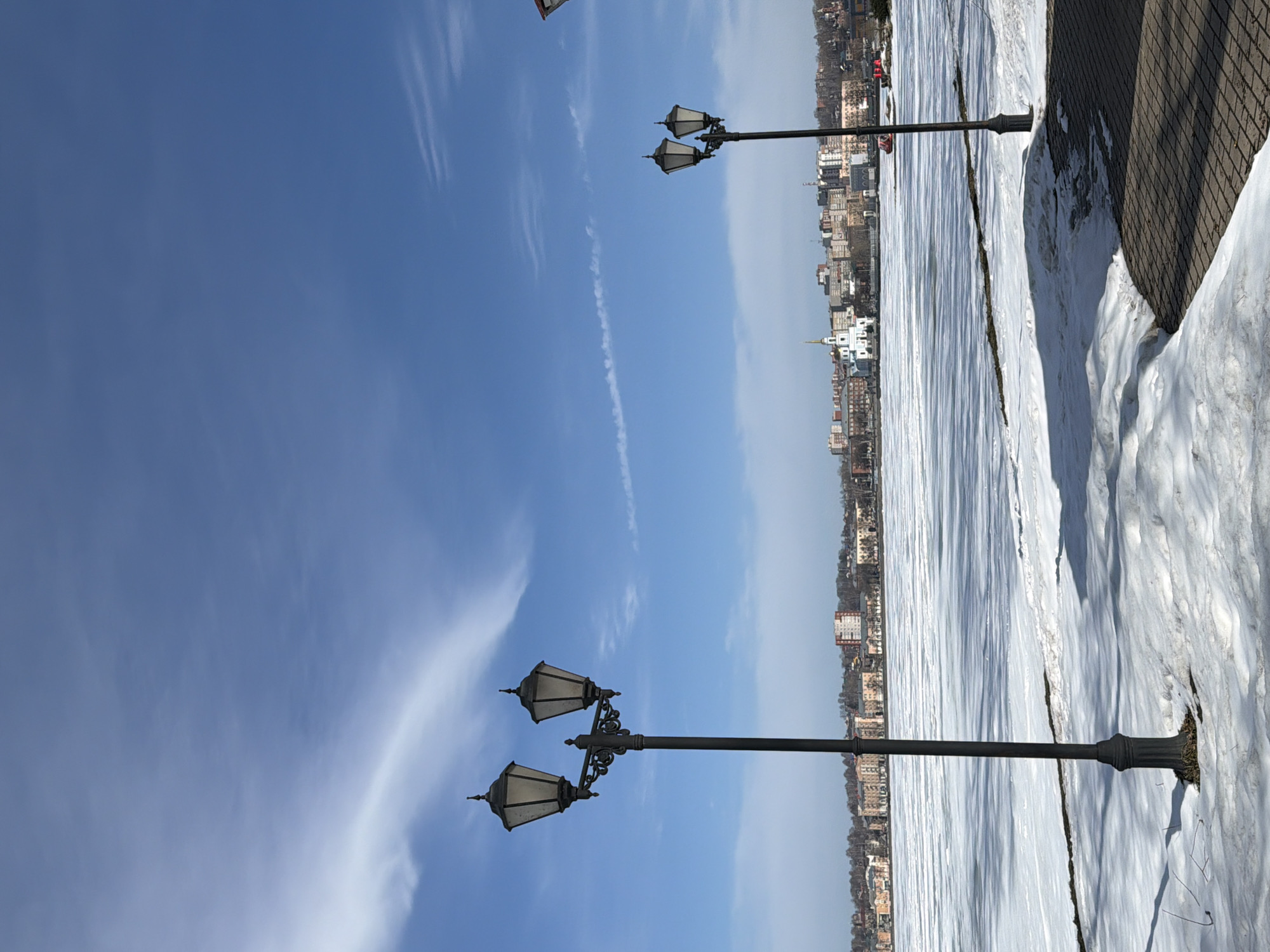 The image depicts a winter scene in Votkinsk, Russia. Two vintage lampposts stand prominently in the foreground against a bright blue sky with scattered cirrus clouds and a contrail. In the midground, the partially frozen Votkinsk Reservoir stretches out toward the horizon, covered in snow and ice. Beyond the reservoir, the city skyline is visible, featuring a mix of low-rise buildings, including the distinctive white and gold spire of the Annunciation Cathedral. A paved walkway borders the frozen reservoir, with snow piled along its edge.