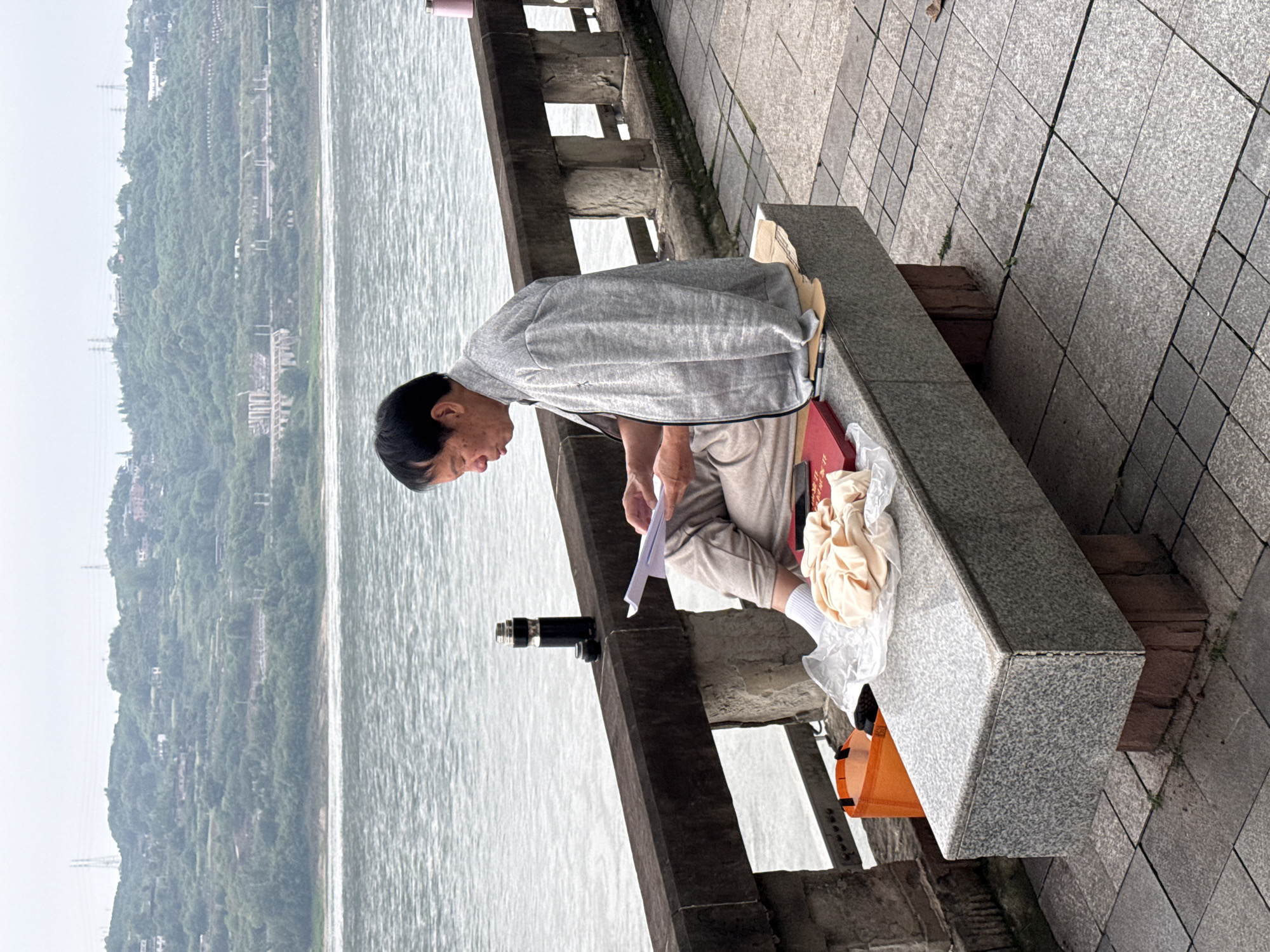 An elderly person sits on a stone bench writing near the riverbank in Jiangjin, China. The person is wearing beige pants, white socks, and a gray zip-up sweater. They are holding a piece of paper. Next to them on the bench is a red folder, a plastic bag filled with clothes, and an orange grocery bag hanging on the bench's side. The bench is part of a stone barrier separating the sidewalk from the river. The sidewalk is paved with gray square tiles. Behind the barrier is a large body of water with a tree-covered hill on the opposite bank.