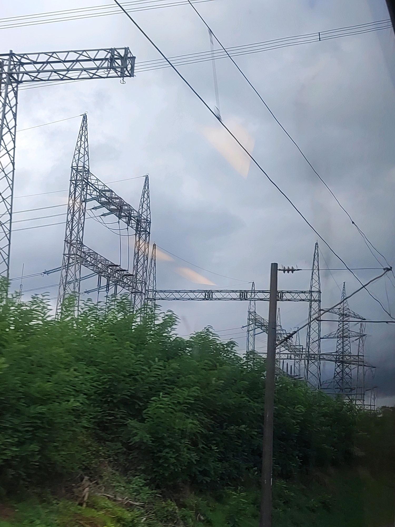 The image shows a power substation with tall steel towers supporting electrical transmission lines against a cloudy sky. The towers are interconnected by a complex network of wires. In the foreground, there is dense green foliage, suggesting a rural or semi-rural setting. The lighting indicates it is daytime, possibly overcast or approaching dusk. There are no people or vehicles visible in the scene. The image appears to be taken from a moving vehicle, as indicated by the slight blur and the reflection on the surface from which the photo was taken. The power substation is likely located in Roßtal, Germany, given the context.