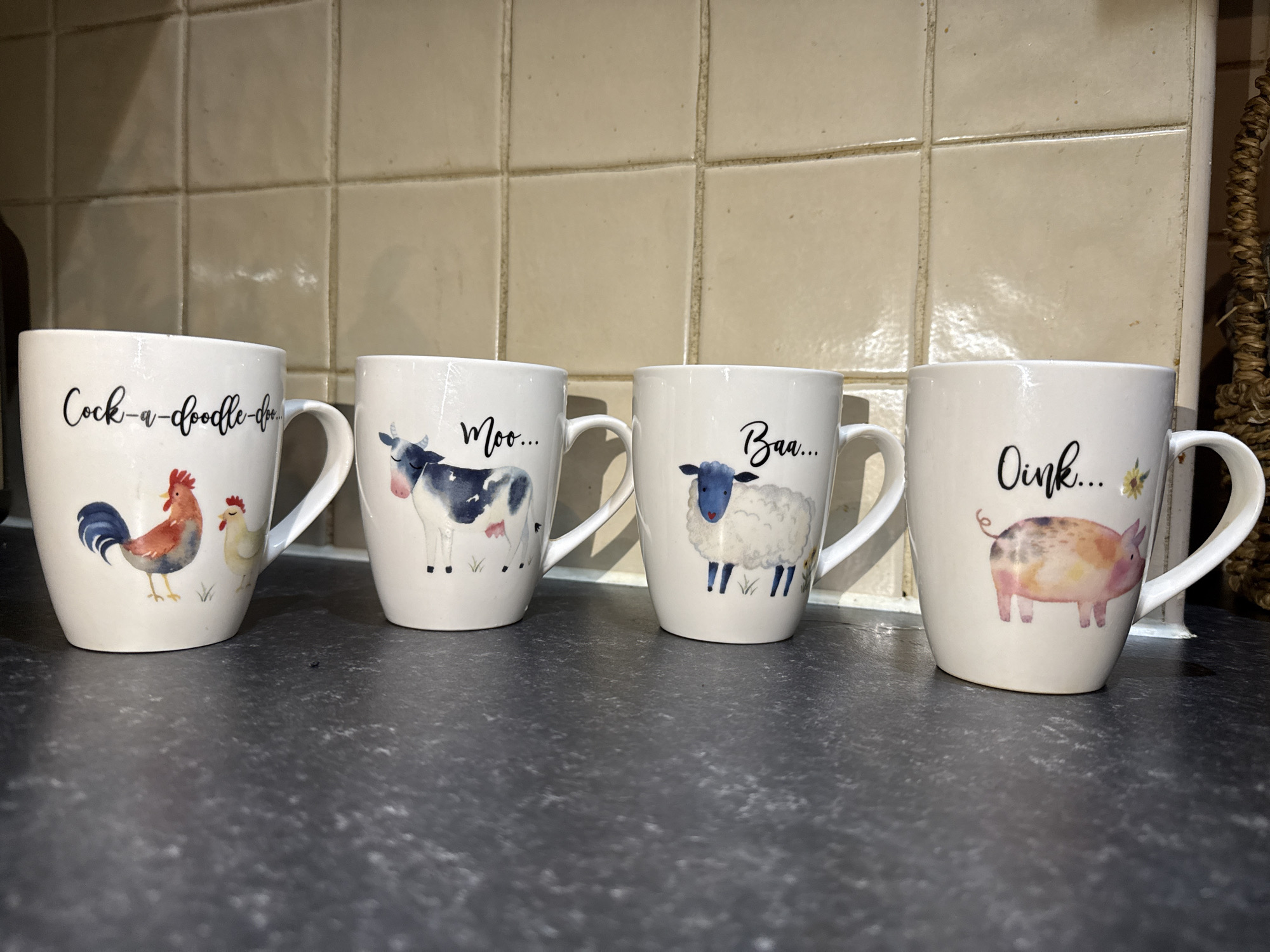 Four white mugs with farm animal images are arranged in a row on a dark countertop. The mugs feature a rooster and hen with the text "Cock-a-doodle-doo...", a cow with "Moo...", a sheep with "Baa...", and a pig with "Oink...". The background is a tiled wall, and a woven basket is visible to the right of the pig mug. The location is not specified in the image.