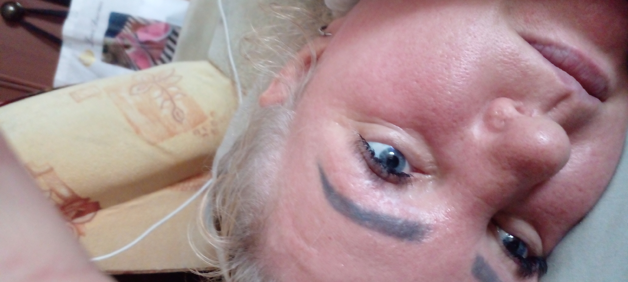 The image is a close-up, slightly angled shot of a woman's face, cropped to focus on her left eye, nose, and mouth. Her blonde hair is visible at the top of the frame, with wisps falling around her face. She has blue eyes, with noticeable eyelashes and well-defined eyebrows. Her skin appears flushed and textured. A hint of her lips is visible, slightly parted. In the background, out of focus, is a light-colored, patterned pillow or fabric, and a glimpse of what appears to be a white bag. There is no visible indication of the city of Glubokoe, Kazakhstan, nor any details to place the image there. The lighting suggests an indoor setting.