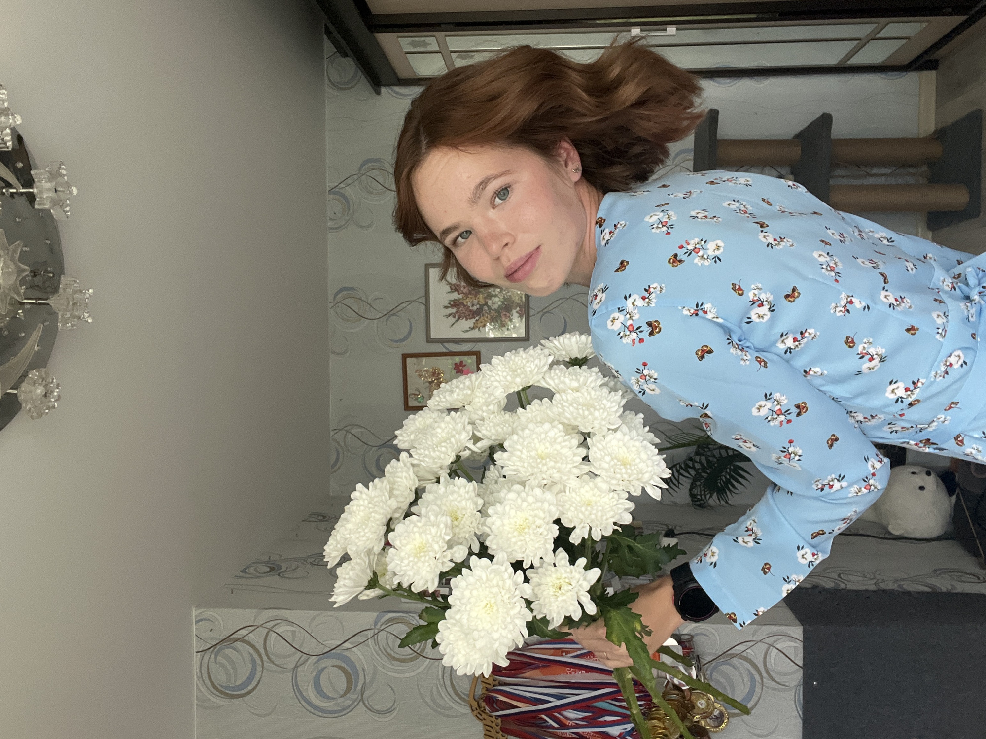 A woman with auburn hair stands in a room, holding a bouquet of white chrysanthemums. She is wearing a light blue dress with a floral and butterfly pattern. The room has patterned wallpaper and a ceiling fixture with crystal accents. In the background, there is a tall closet, a cat tree, and a framed picture on the wall. The woman is positioned slightly off-center, looking over her shoulder towards the camera. The lighting suggests an indoor setting during the daytime. The location is identified as Chaikovsky, Russia.