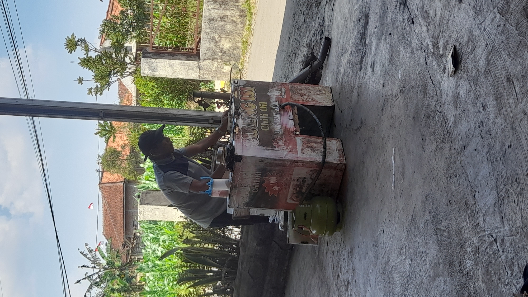 The image depicts a street vendor preparing food at a roadside stall in Selaawi, Indonesia. The vendor, a man wearing a grey t-shirt and a baseball cap, is focused on his work at a mobile cart. The cart is decorated with signage that reads "CIRENG ISI Chili Oil" and "ENAK LEZAT HALAL," indicating the food being sold is likely chili-filled cireng, a type of Indonesian snack. A green gas cylinder sits to the left of the cart, connected by a hose. In the background, a residential area with traditional Indonesian houses with red tiled roofs is visible, along with lush green vegetation, including corn crops and trees. The ground is a textured, grey, concrete surface. The overall scene suggests a typical day in a semi-urban or rural area of Indonesia.