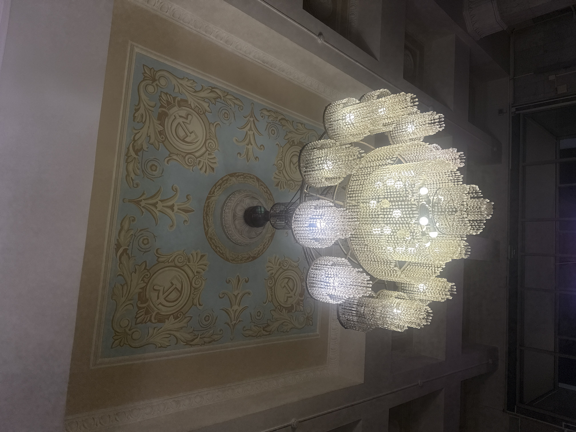 The image depicts a chandelier and ceiling in what appears to be a large, possibly ornate room. The chandelier is multi-tiered, with numerous crystals creating a sparkling effect. The ceiling has an intricate design featuring what seems to be the hammer and sickle emblem, surrounded by decorative patterns in light blue, gold, and beige tones. This pattern is likely part of the historical architecture. Considering the hammer and sickle imagery, and given the question's reference to Yekaterinburg, Russia, this image is highly suggestive of a location built during the Soviet era. Without other contextual clues, it is challenging to specify the precise location, but potential candidates may include government buildings, palaces of culture, or major public spaces that would reflect the city's historical significance. The room itself looks to be a historical ballroom or an old Soviet auditorium.