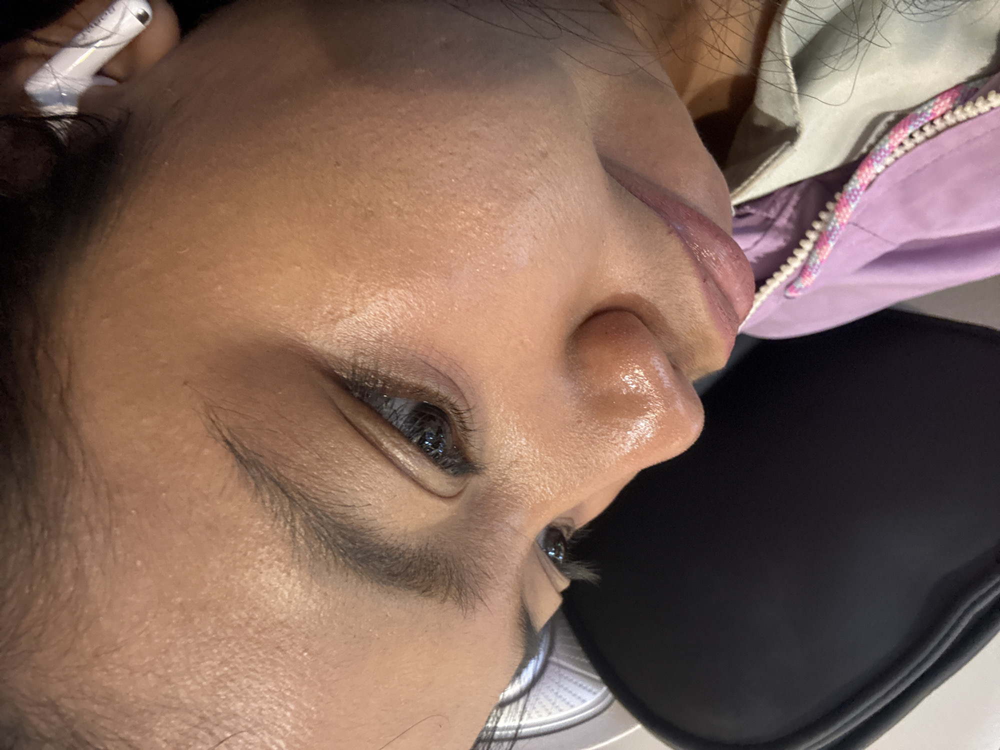 The image shows a close-up of a person's face, tilted upwards and to the right, resting against a dark headrest. The person appears to be indoors, possibly in a vehicle or waiting area, given the context of the headrest and what appears to be the edge of a seat. The person's skin is visible, with some slight sheen, suggesting it might be humid or the person has been active. The person is wearing a light purple jacket with a colorful zipper pull. A white earbud is visible in the person's right ear. The lighting is soft and diffused. No specific geographical markers are present to indicate a location within North Jakarta, Indonesia.