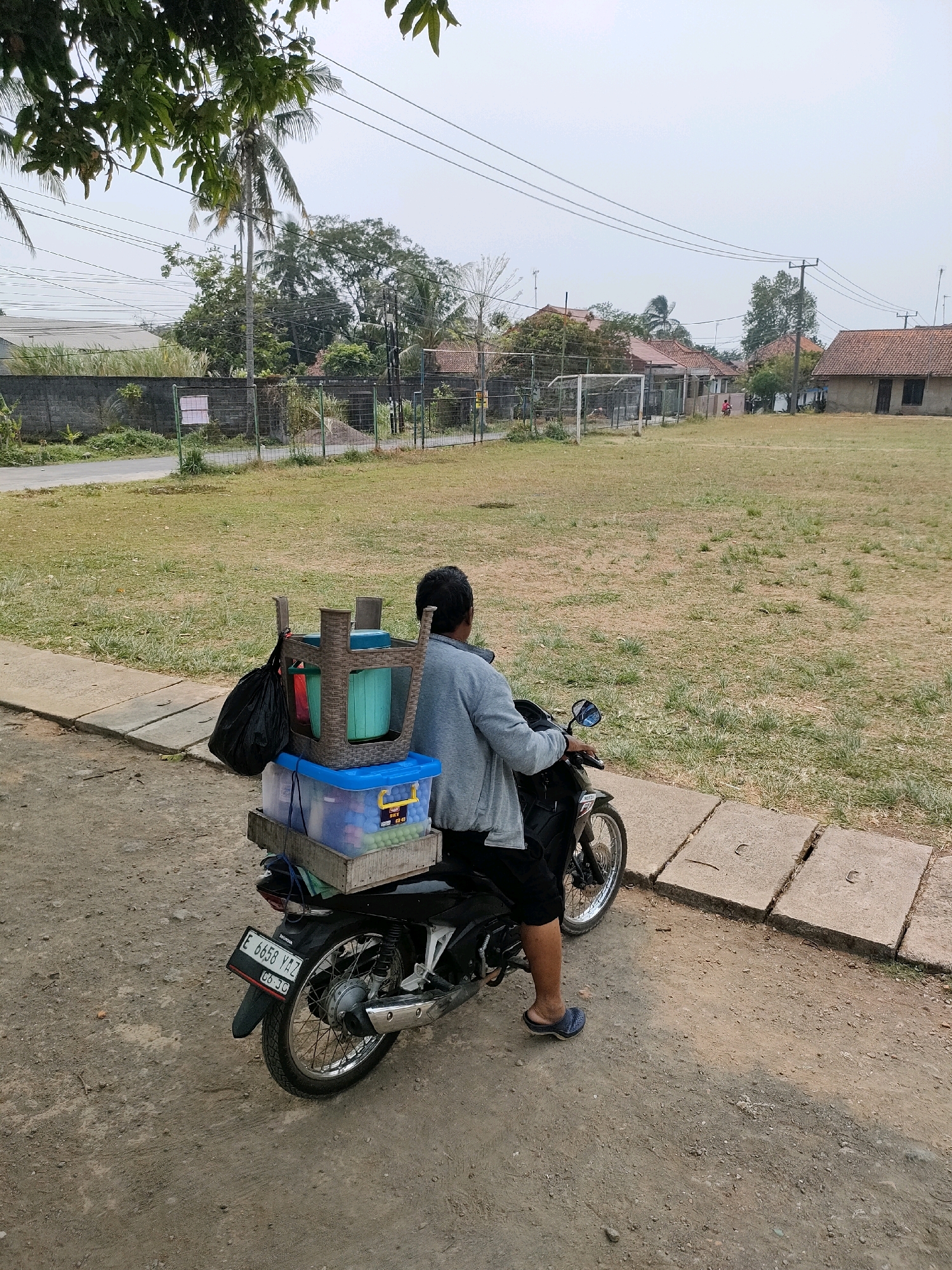 A person sits on a black motorcycle, facing away from the camera, parked on a dirt road next to a paved sidewalk. The motorcycle is laden with several items on its rear rack. Stacked on the rack are a blue plastic bin containing small, colorful balls, a wooden crate, a blue bucket, and a black plastic bag. A brown wicker basket-like object sits atop the blue bucket, holding what appears to be more buckets or containers and a red item. The person is wearing a grey long-sleeved shirt, black shorts, and blue slip-on shoes. The background depicts a grassy field, a fence, and several buildings with tiled roofs, suggesting a residential area. A soccer goal is visible in the distance on the left side of the field. The sky is overcast. The location is identified as Bojong, Indonesia.