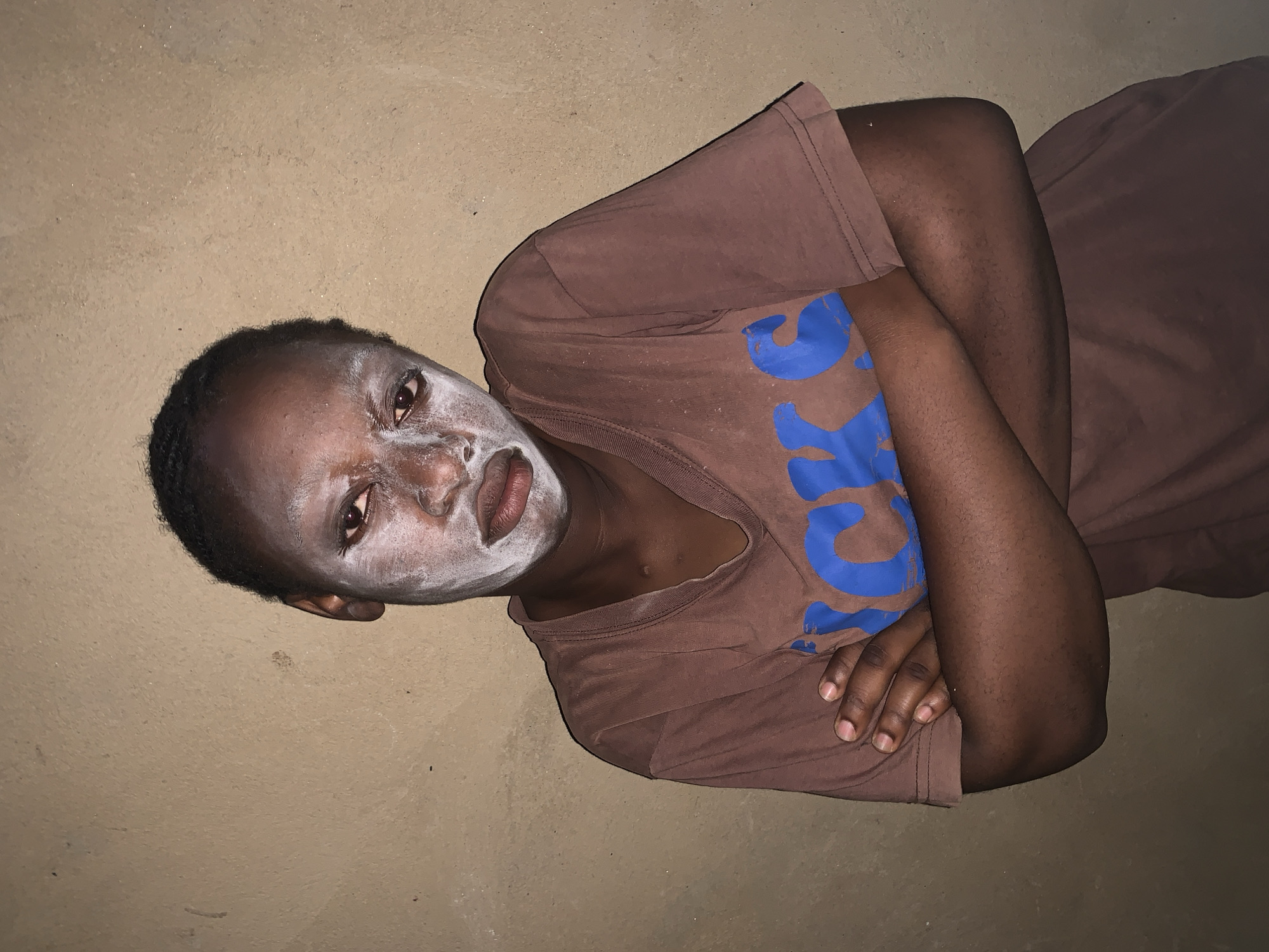 A person with dark skin and short, dark hair is lying on what appears to be a tan or light brown surface, possibly a floor or bed. The person's face is covered in a white powdery substance, possibly makeup or a mask. They are wearing a brown t-shirt with faded blue lettering. Their arms are crossed over their chest. The setting is unclear but described as Agban, Nigeria. The lighting is dim.