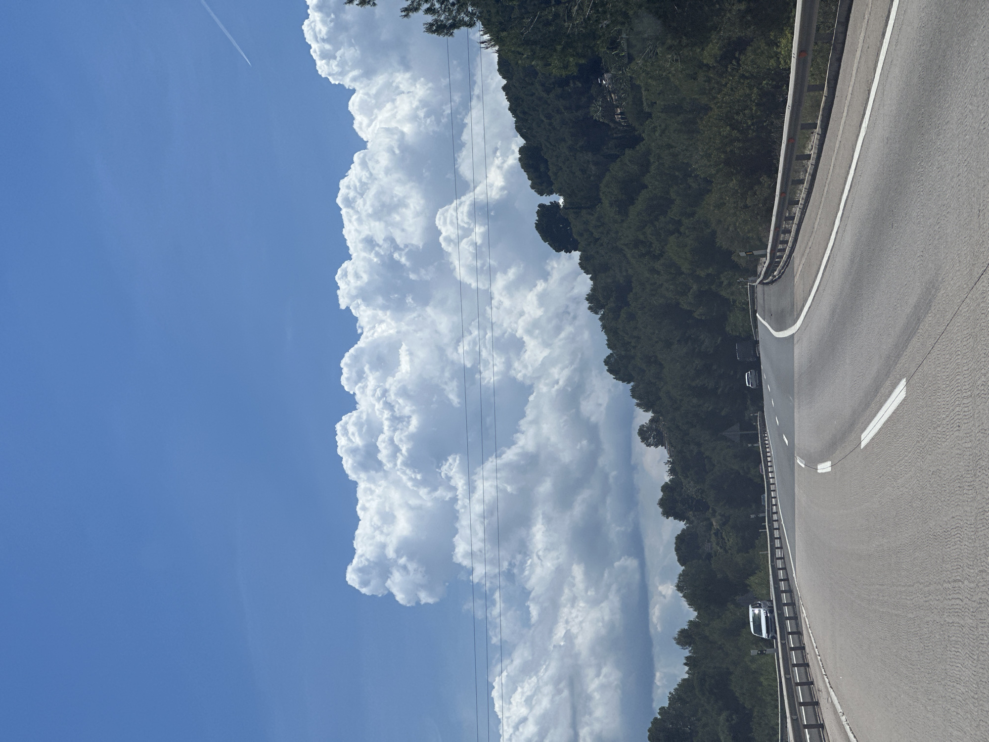 A low-angle shot depicts a highway scene likely near la Font d'en Carròs, Spain, under a bright blue sky featuring large cumulus clouds. The right side of the frame shows a highway with a guardrail and white lane markings. A line of trees runs parallel to the highway on the left. Two cars can be seen on the highway. There are power lines crossing over the view of the clouds. A jet contrail is visible in the blue sky at the top left of the image.