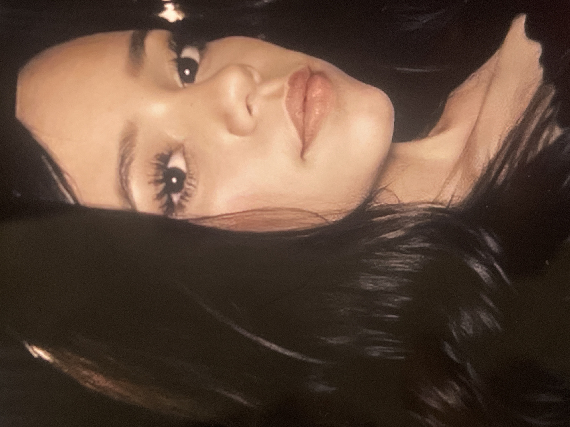 The media file displays a close-up portrait of an individual, likely a female, positioned against a dark background, possibly composed of their own long, dark hair. The subject's head is tilted, suggesting they are reclining or posing. The image primarily focuses on their face, from the nose upwards, with a portion of their neck and collarbone visible on the right side of the frame.
The person exhibits a light skin tone, and their eyes are open, with a direct gaze towards the viewer. Distinctive features include long, defined eyelashes and well-shaped eyebrows. Their lips appear full, glossy, and are adorned with a neutral or light pink lip product. The nose is clearly visible. Soft, diffused lighting illuminates the face, emphasizing its contours and the texture of the skin. The dark hair frames the face and blends into the background, creating a strong contrast with the skin.
No specific activity is depicted beyond posing for the photograph. The expression conveyed is serene or contemplative. There are no visible objects other than the person and their hair, nor any discernible setting cues beyond the dark background. No text is present in the image. The time of day and weather cannot be determined from the photograph.