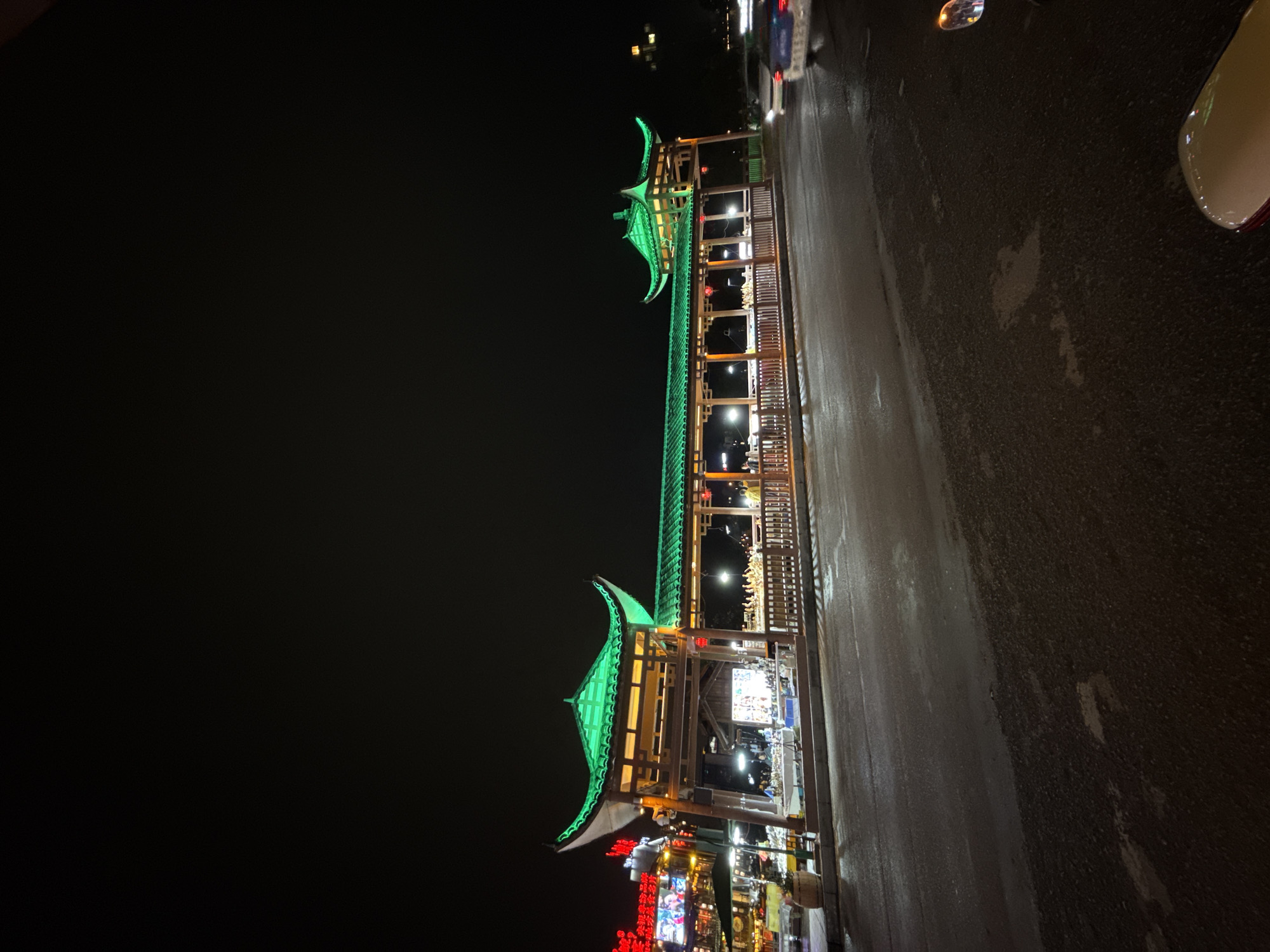The image captures an illuminated traditional Chinese architectural structure, likely a gateway or pavilion, in Wulingyuan, China. The structure features a multi-tiered roof design with upturned eaves, adorned with vibrant green lighting. Ornate wooden framework supports the roof, and the spaces between the roof tiers are filled with decorative elements and illuminated fixtures. Below the structure, a variety of neon signs and lights indicate the presence of shops or commercial establishments. The surrounding environment appears dark, suggesting it is nighttime. The foreground shows a paved surface, potentially a street or walkway, with some reflective patches that might indicate moisture. A portion of a vehicle is visible on the right side of the frame. There are no people visible in the immediate foreground.