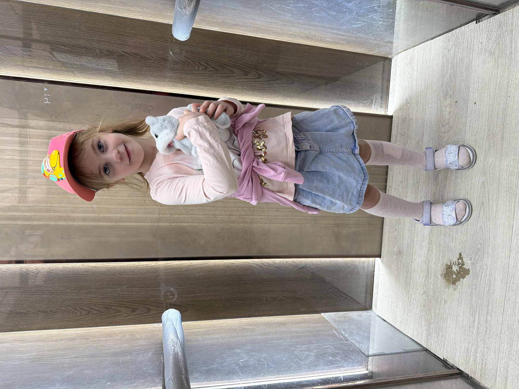 The image depicts a young girl standing in what appears to be an elevator. She is holding a stuffed animal and wearing a pink baseball cap with a cartoon character, a light pink long-sleeved shirt, a light blue denim skirt, white knee-high socks, and light purple sandals. A pink sweatshirt is tied around her waist. The elevator walls are paneled with a wood-like material and have vertical lighting elements. The floor is light-colored and appears to have some moisture or a stain. The location is described as being in the city of Perm, Russia.