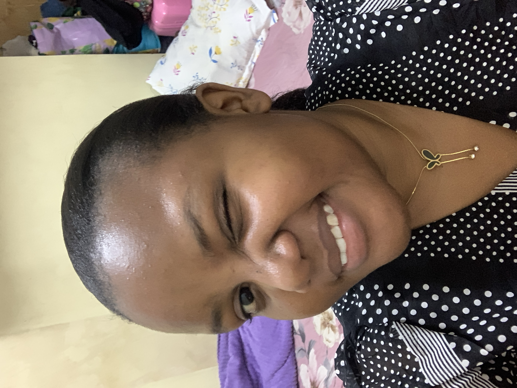 A close-up indoor shot depicts a dark-skinned woman smiling with her right eye slightly closed. She has black hair pulled back from her forehead. The background includes a light yellow wall, a pillow with a floral pattern, a pink blanket, and a purple cloth. The woman is wearing a black dress with white polka dots and stripes, and a gold necklace with a butterfly-shaped pendant. The location is unknown, and there is no evidence to indicate that this photo was taken in Dar es Salaam, Tanzania.