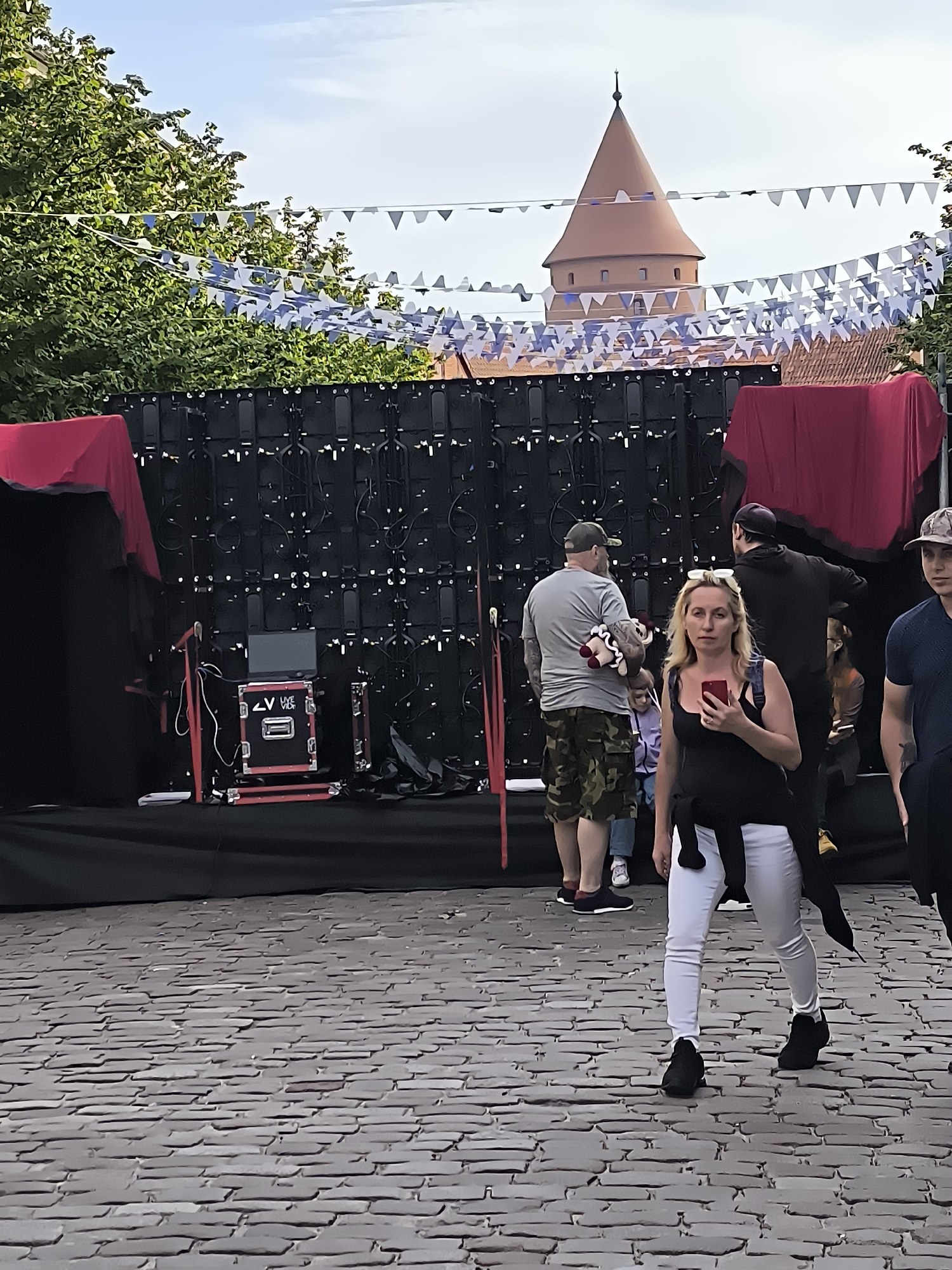 The image depicts an outdoor scene in Klaipėda, Lithuania, with a prominent brick-paved area in the foreground and a large LED screen setup in the background. Several people are present, engaged in various activities. A woman in the foreground is holding a red smartphone and looking towards the camera. To her left, a man in camouflage shorts and a grey t-shirt is holding a stuffed animal. Behind them, more people are visible, and a conical tower, possibly part of a historical building, is seen in the distance, along with festive pennant banners strung across the sky. The background also includes what appears to be a stage setup with red drapery and various technical equipment. The overall impression is that of a public event or gathering.