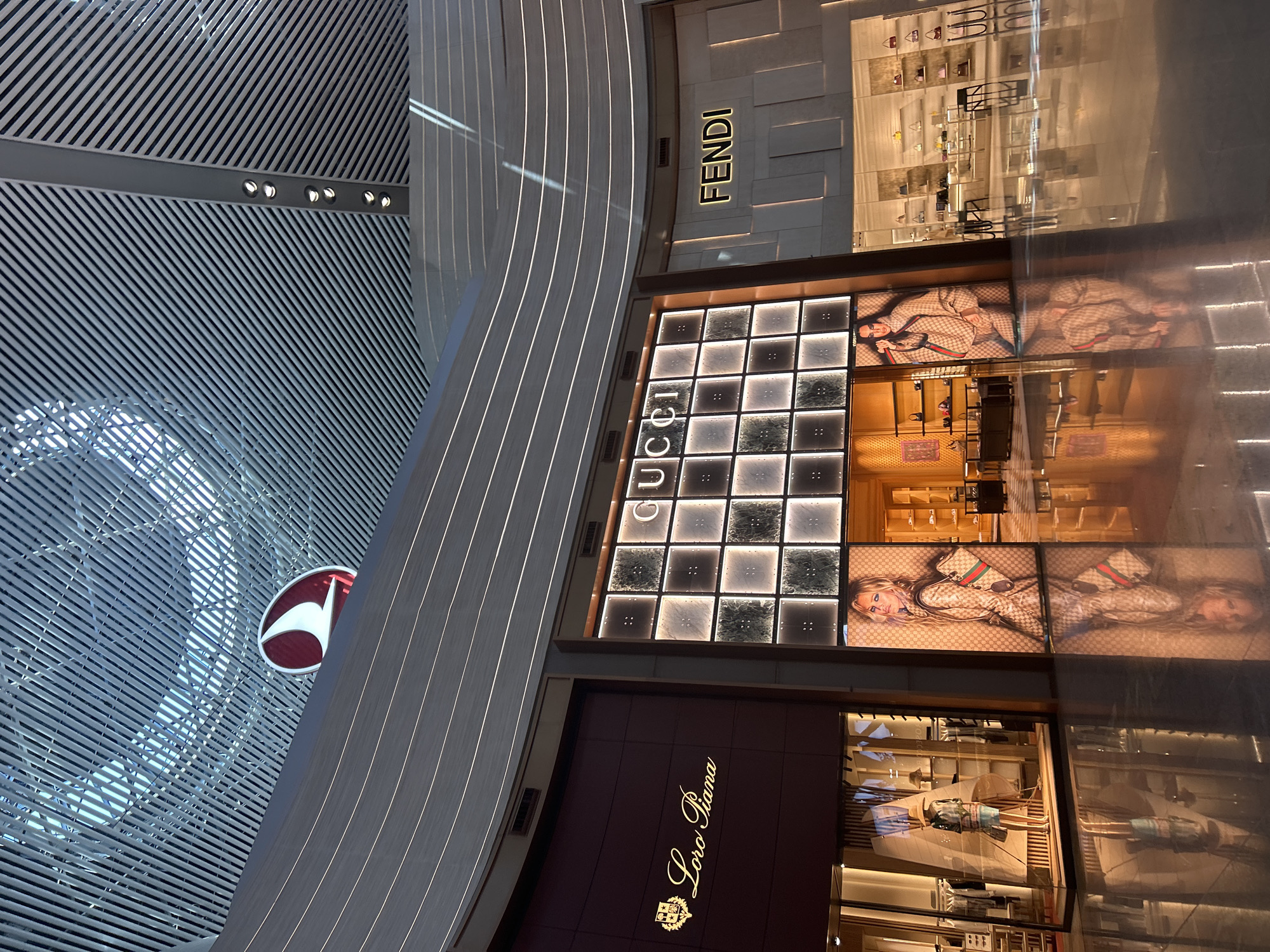 This media file captures a brightly lit, modern indoor commercial environment, likely a luxury shopping concourse within an airport. The scene is dominated by the illuminated facades of high-end retail stores, viewed from an upward and oblique angle.
Prominently featured are three luxury brand stores: Fendi, Gucci, and Loro Piana. The FENDI store, located at the top right, displays its name in gold lettering against a textured, light-toned wall. Below Fendi, the GUCCI store entrance is a grid of illuminated square panels, with "GUCCI" written vertically in bold letters on the left. Large display advertisements within the Gucci windows show models dressed in branded attire, with reflections creating multiple instances of the imagery. Below Gucci, the Loro Piana store is identifiable by its elegant gold script "Loro Piana" and crest logo against a dark, rich-toned facade. Merchandise and mannequins are visible through the store windows.
The architectural setting includes a distinctive, intricately patterned ceiling or wall on the left, composed of numerous parallel and diagonal metallic slats. Integrated strip lighting runs along curved structural elements, contributing to the modern aesthetic. On the far left, partially obscured, is a red circular logo with white elements, which is the identifiable emblem of Turkish Airlines, strongly indicating an airport location. The strong artificial lighting and reflections suggest an indoor environment, likely during evening hours or at night. The overall atmosphere is one of upscale commerce and contemporary design.