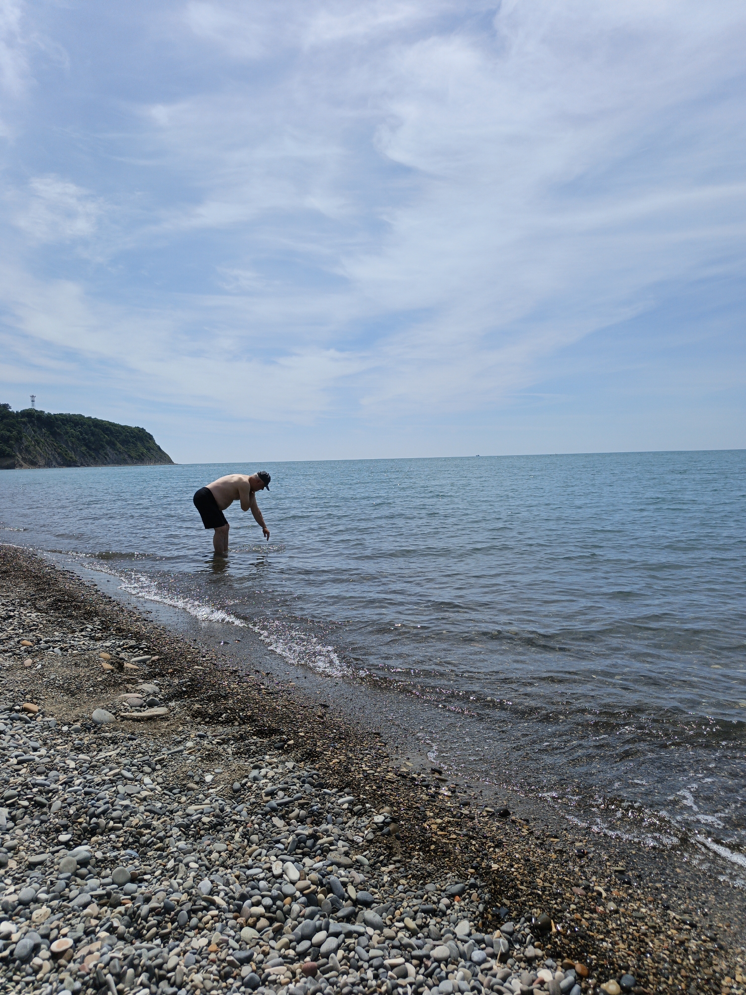 The image presents a coastal scene in Krinitsa, Russia, under a partly cloudy sky. In the midground, a lone adult male is standing in the shallow, calm seawater, bent at the waist with his right hand submerged just below the surface. He is shirtless, wearing dark shorts and a dark baseball cap. The foreground reveals a pebble beach composed of numerous rounded stones in shades of grey, brown, and white, extending from the bottom of the frame to the water's edge. Small waves gently break on the shore, creating minor foam and glints of light on the water's surface. The sea, exhibiting a blue-grey hue, stretches towards the horizon with a placid surface. On the left side of the frame, a verdant, forested hill or cliff slopes down to meet the water. A tall, slender tower-like structure is visible atop this landmass. The sky occupies the upper half of the image, featuring scattered, wispy white clouds against a pale blue backdrop.
