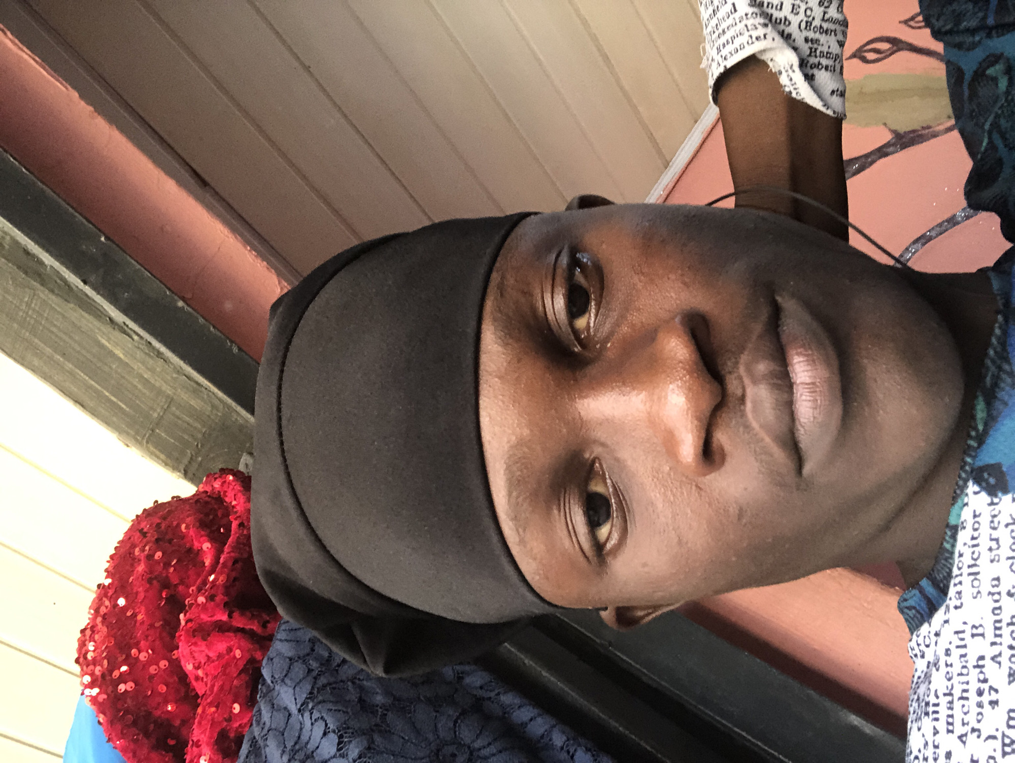 This is a close-up selfie of a young Black male wearing a black kufi. He is positioned in the center of the frame, with his face taking up most of the image. The background consists of wooden planks, some painted in a peachy-pink color, and other dark, possibly metal, beams. To the left, there are hints of fabric, including a bright red material covered in sequins and a dark blue lace fabric. The lighting appears to be natural, possibly from an overhead source, casting a warm glow on his skin. The image is an outdoor shot, likely taken in a relaxed, informal setting. There is no specific identifiable landmark or indication of a location within Ado, Nigeria, beyond the general appearance of the background structures which are common in many regions.