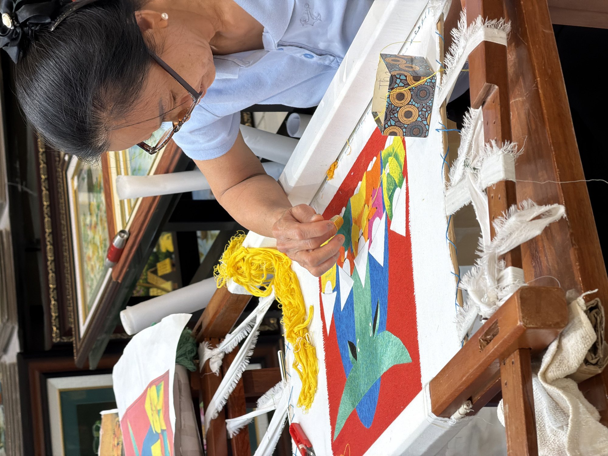 An older woman, likely in Hải Phòng, Vietnam, is deeply focused on crafting a vibrant textile piece, possibly a rug or tapestry, stretched on a large wooden frame. She is positioned to the right of the frame, leaning over her work, with her hands manipulating colorful yarn or thread. Her dark hair is pulled back into a neat bun, adorned with a black scrunchie and a decorative silver and pearl clip. She wears brown-rimmed glasses and a light blue polo shirt with a small anchor emblem embroidered on the chest.
The textile displays an abstract, geometric design rendered in bright primary and secondary colors including red, yellow, green, and blue, contrasting with white sections. A prominent tangle of bright yellow yarn sits near her left hand, suggesting the current color she is using or has recently used. A small red tool, possibly scissors or a cutter, is partially visible near the lower right edge of the textile. The sturdy wooden frame holding the artwork is wrapped in various pieces of white fabric, some frayed, securing the textile in place. A small box with a yellow and blue geometric pattern is attached to the upper part of the frame with yellow string.
In the background, out of focus, are multiple framed pictures or paintings and white rolled items, likely canvases or prints. This suggests the setting is an art studio, workshop, or a section of a gallery. The woman appears absorbed and dedicated to her intricate craft. No discernible text is visible in the image.