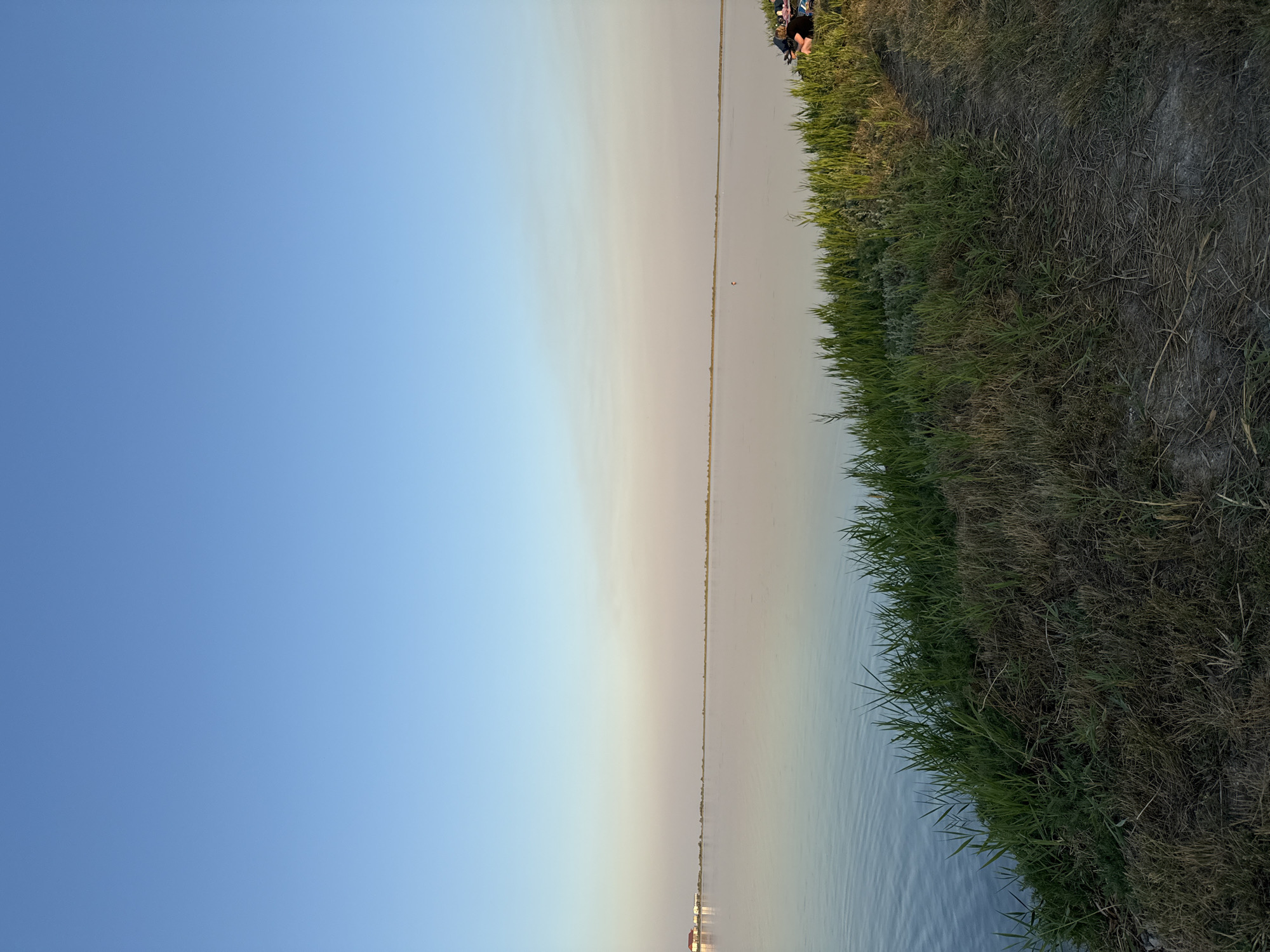 In Serhiivka, Ukraine, a tranquil scene unfolds by the water. The image captures a wide expanse of calm water reflecting the clear blue sky. The horizon is marked by a distant, low-lying shoreline. In the foreground, a grassy embankment slopes down to the water's edge. The grass is lush and green in some areas, transitioning to dry, brown vegetation in others. A dirt path is visible on the embankment, leading towards the right side of the frame.
On the right side of the image, near the top of the embankment, two individuals are seated on chairs. They appear to be relaxed, with one person looking towards the water. Several items, including bags and possibly blankets, are arranged around them. A small orange buoy is visible in the water, further out from the shore. In the distance, along the left-hand horizon, a building with a red roof is faintly discernible. The overall atmosphere is peaceful and serene, suggesting a leisurely day by the water.