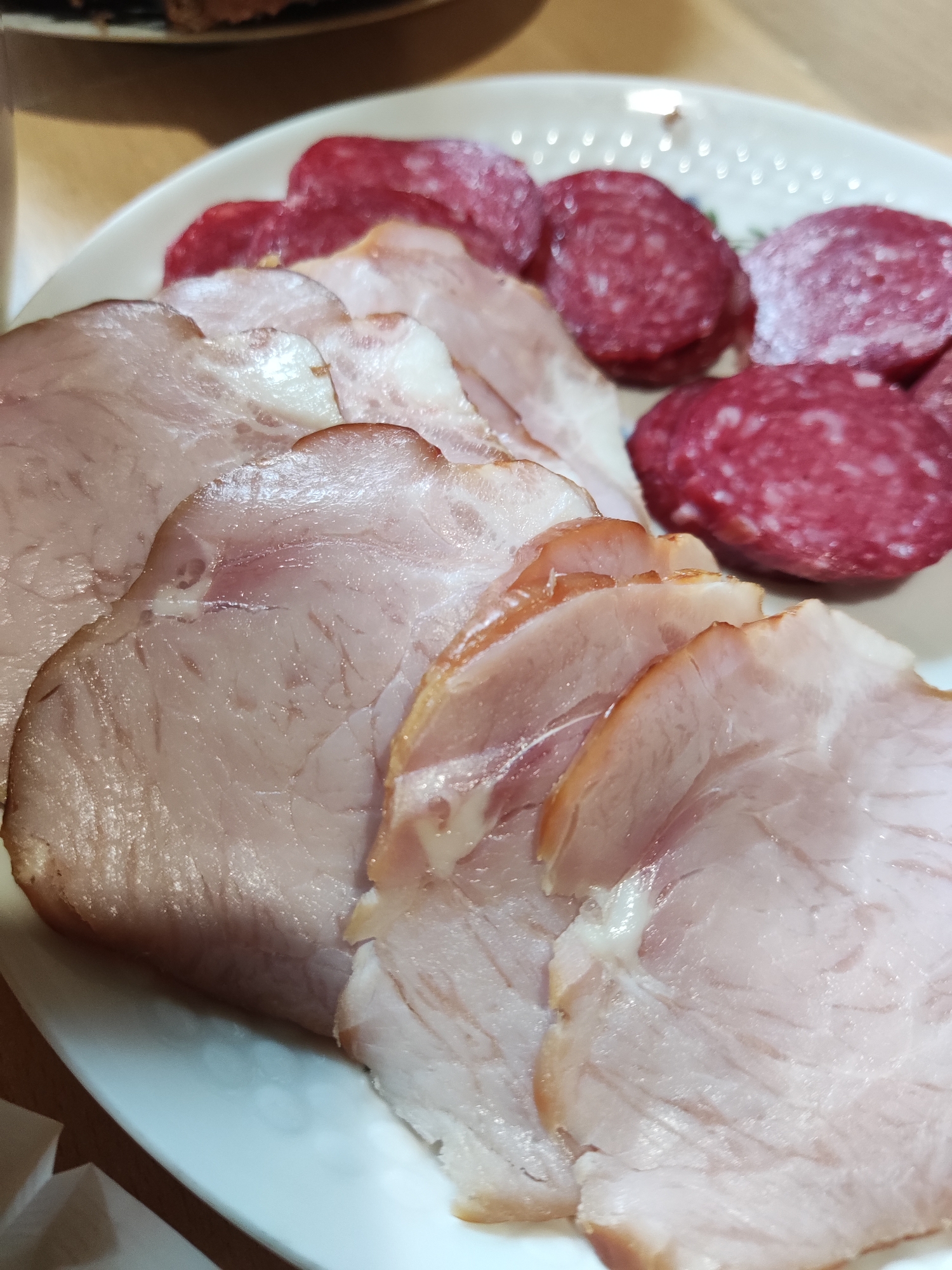 A close-up, overhead shot of sliced ham and salami on a white plate. The ham slices are arranged in the foreground, with some overlapping. They are light pink and have visible fat marbling. Behind the ham, slices of red salami are visible, with visible white fat speckles. The plate has a decorative rim with small, raised dots. The background is out of focus, but appears to be a wooden table surface. There is no indication of the specific location within Kryvyi Rih, Ukraine.