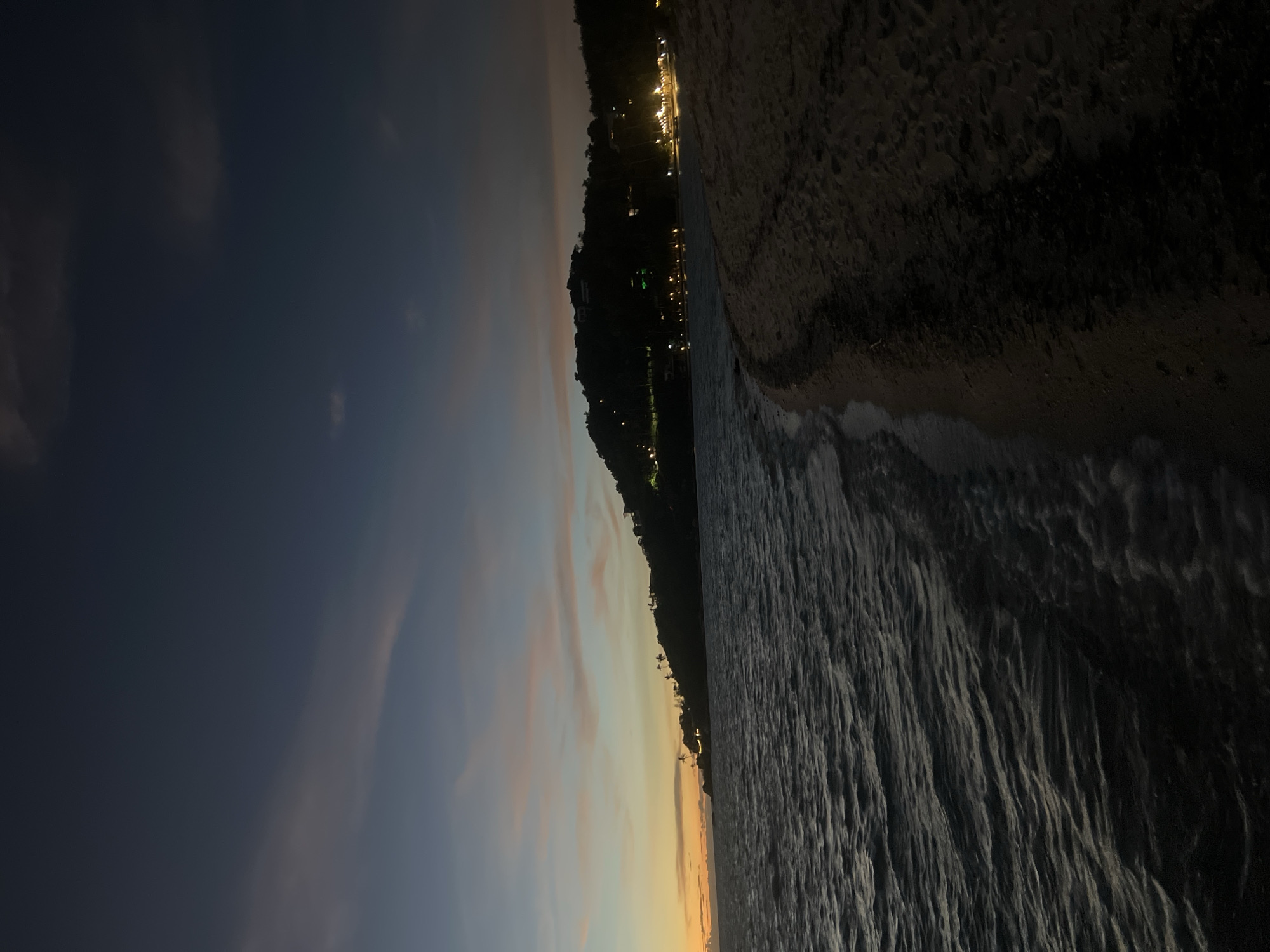 This image depicts a twilight view of a beach on Koh Phangan island, Thailand. The sky is a deep navy blue with streaks of pale pink and orange clouds, indicating the sun has recently set. The ocean is dark and slightly choppy, with gentle waves lapping at the sandy shore. Along the coastline, silhouetted palm trees are visible on a dark, hilly landscape dotted with numerous small, warm-toned lights, suggesting buildings or resorts. The foreground is dominated by the dark sand of the beach, with subtle footprints visible. The overall impression is one of tranquility and the fading light of day.