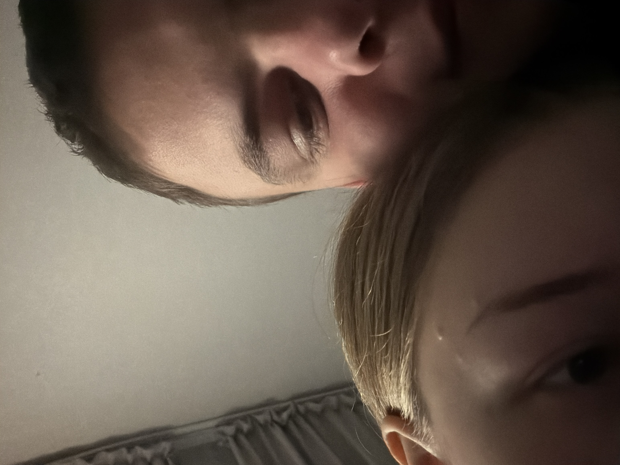 The image depicts a close-up, angled view of two people. One person, with darker hair, is positioned at the top of the frame with part of their face visible, including the nose, an eye, and an eyebrow. The second person, with lighter, blonde hair, is positioned below, with part of their face visible, including an ear, an eye, and an eyebrow. The background appears to be a plain ceiling or wall, with a draped fabric visible at the bottom left. The photograph does not contain any information to ascertain its location as being in the city of Новинки, Russia.