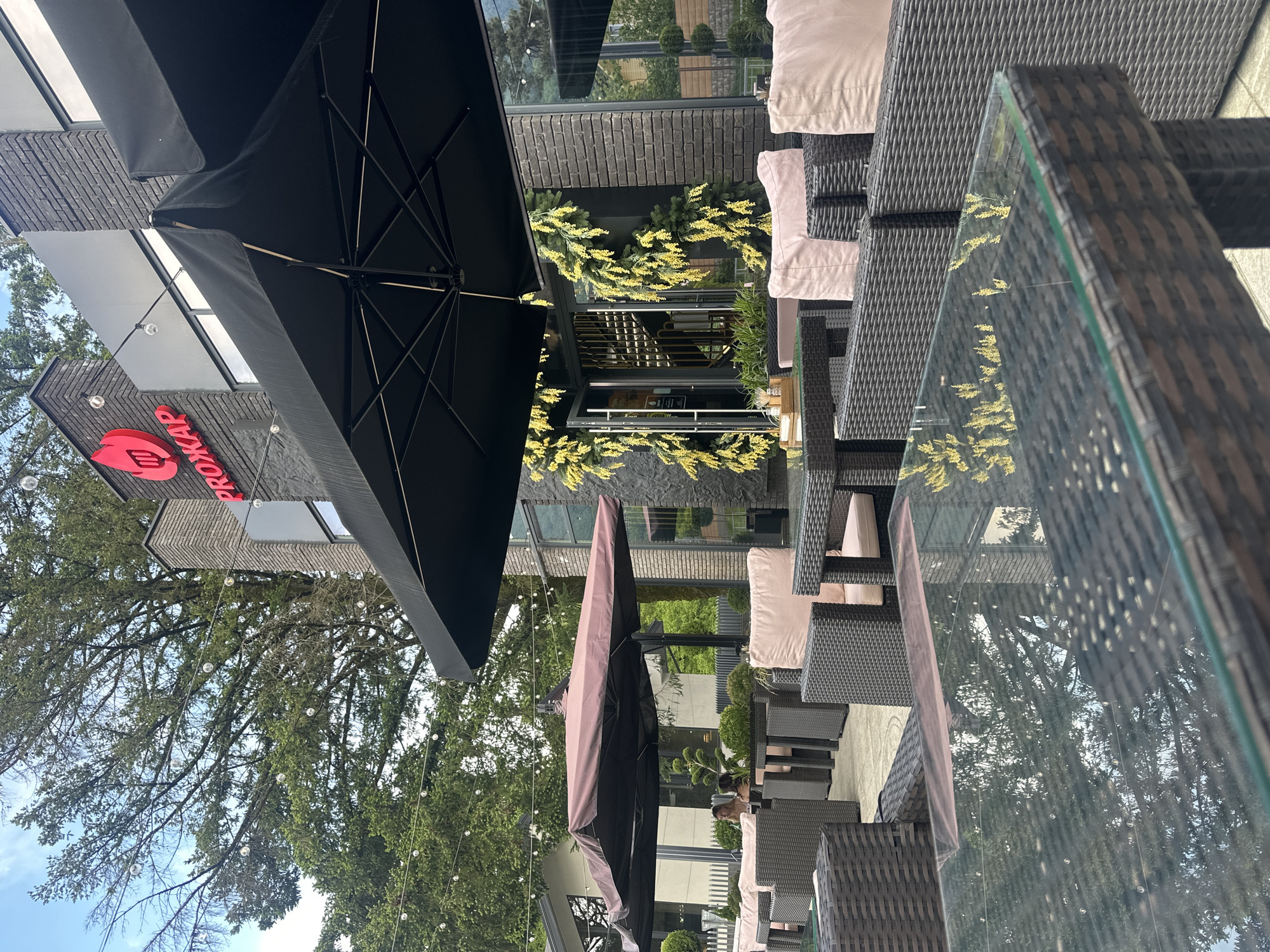 This image captures an outdoor patio dining area of a restaurant named "PROZHAR" (ПРОЖАР) located in Krasnaya Polyana, Russia.
The restaurant building features a dark gray brick facade with large windows. A red sign displaying the restaurant's name in Cyrillic, along with a logo depicting a stylized flame incorporating cutlery, is mounted on the building's upper section. The main entrance is adorned with vertical garlands of yellow foliage.
The patio is furnished with numerous dark brown wicker chairs and tables, each chair equipped with a light pink cushion. The tables have clear glass tops, reflecting the surrounding trees and string lights. Two large, cantilevered outdoor umbrellas provide shade: one black, positioned prominently in the foreground, and another dark brown/maroon umbrella situated further back on the patio. String lights with clear spherical bulbs are strung across the patio overhead.
In the mid-ground, under the dark brown umbrella, two women are seated at a table, appearing to engage in conversation. One woman has dark hair and is wearing a dark top, while the other has lighter hair. Small potted plants or topiaries are interspersed among the tables.
The background reveals lush green trees under a partly cloudy blue sky, suggesting a natural or park-like environment surrounding the restaurant. The ground of the patio appears to be paved with light-colored flagstones or concrete.